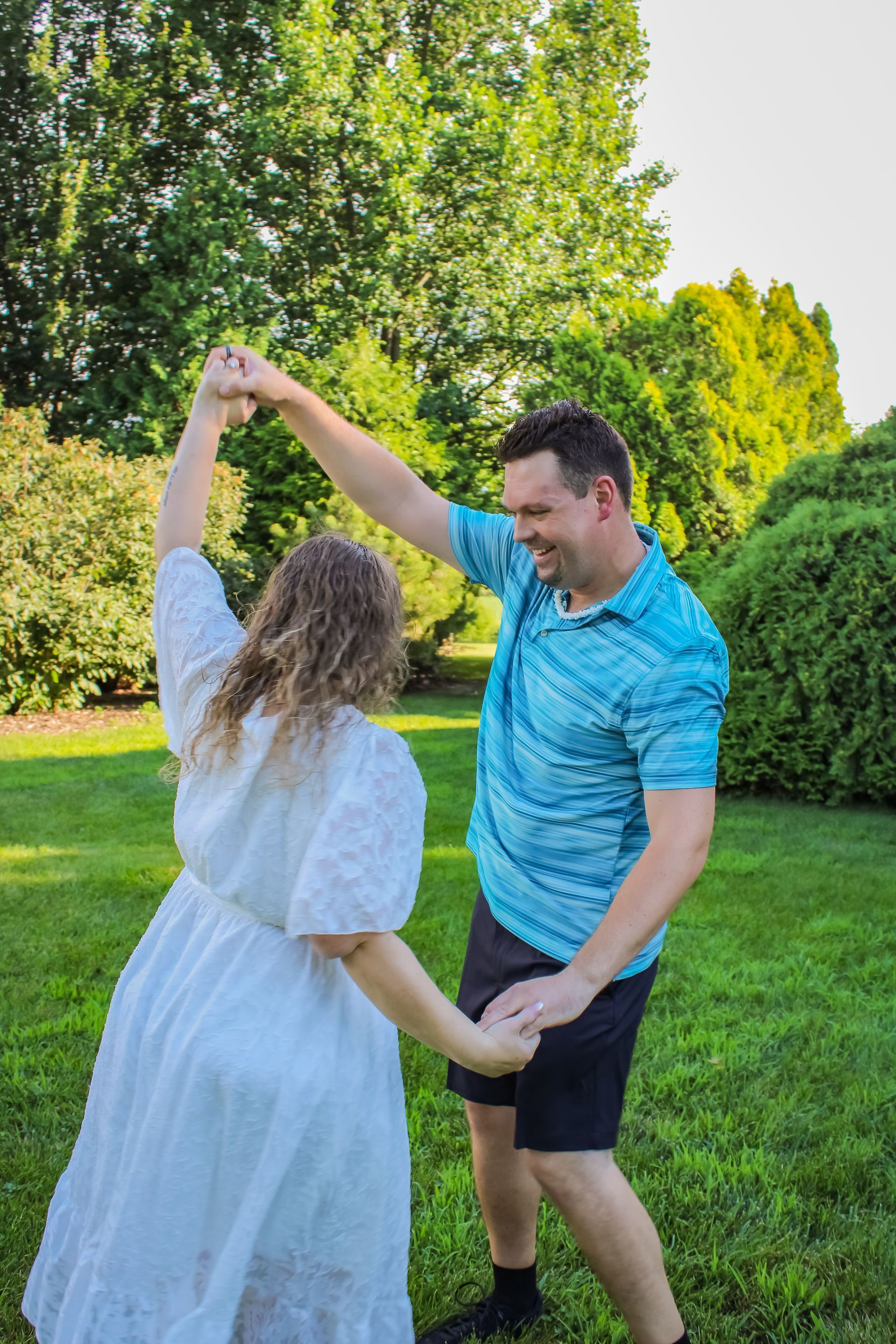 Couple dancing in a sunlit park, woman in white dress, man in blue shirt, holding hands, smiling.