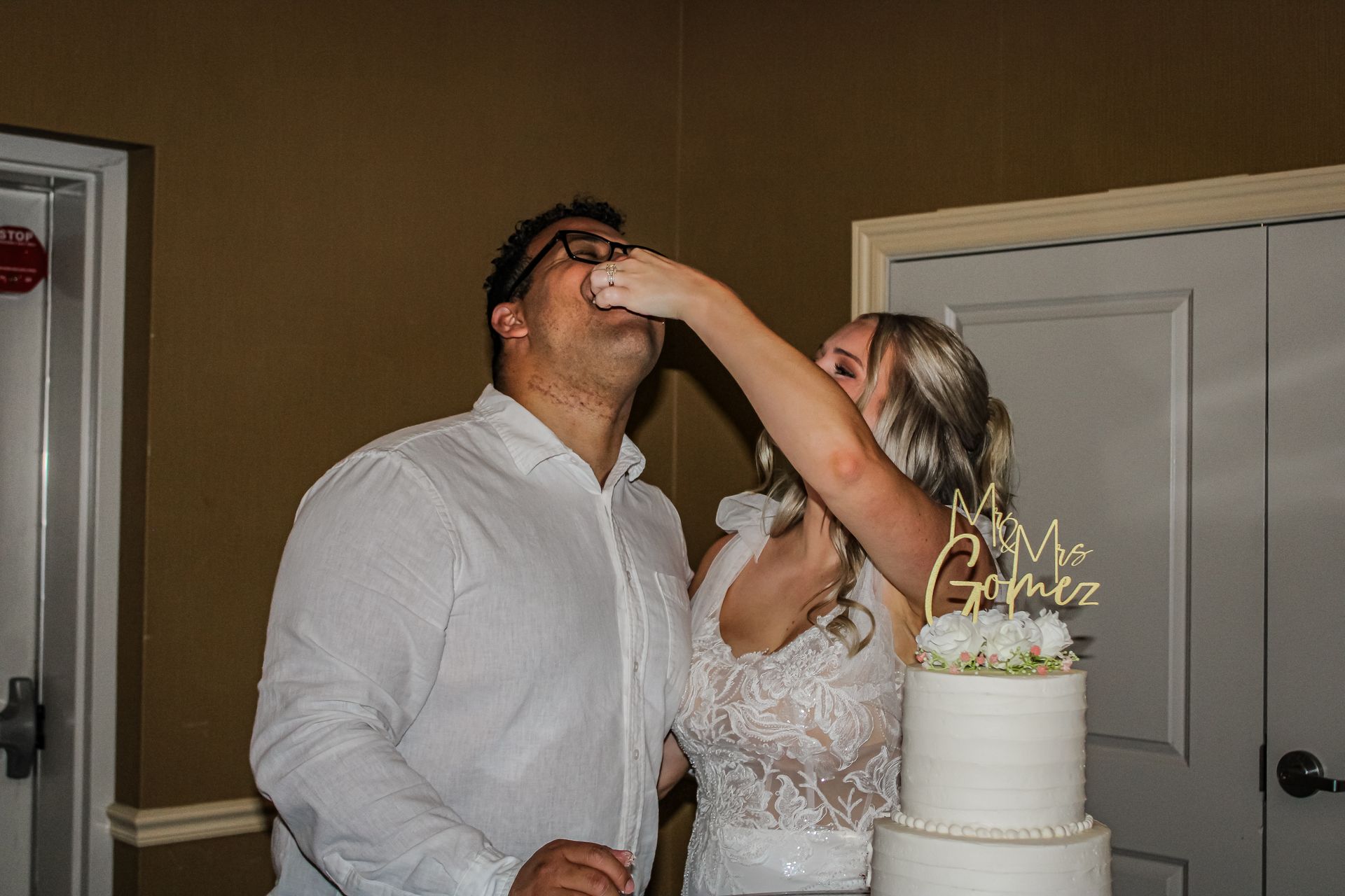 Bride feeding groom wedding cake; couple smiling, celebrating in front of a white tiered cake.