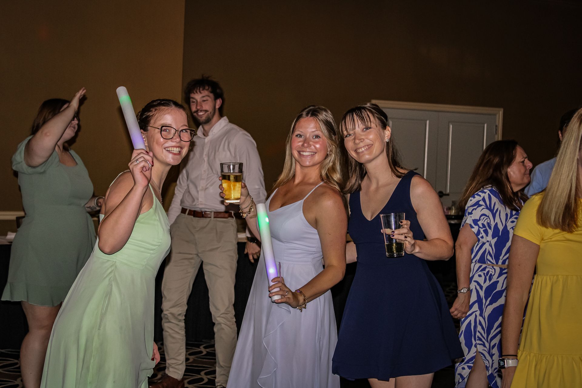 People at a party, some holding glow sticks and drinks, smiling. Dark interior.