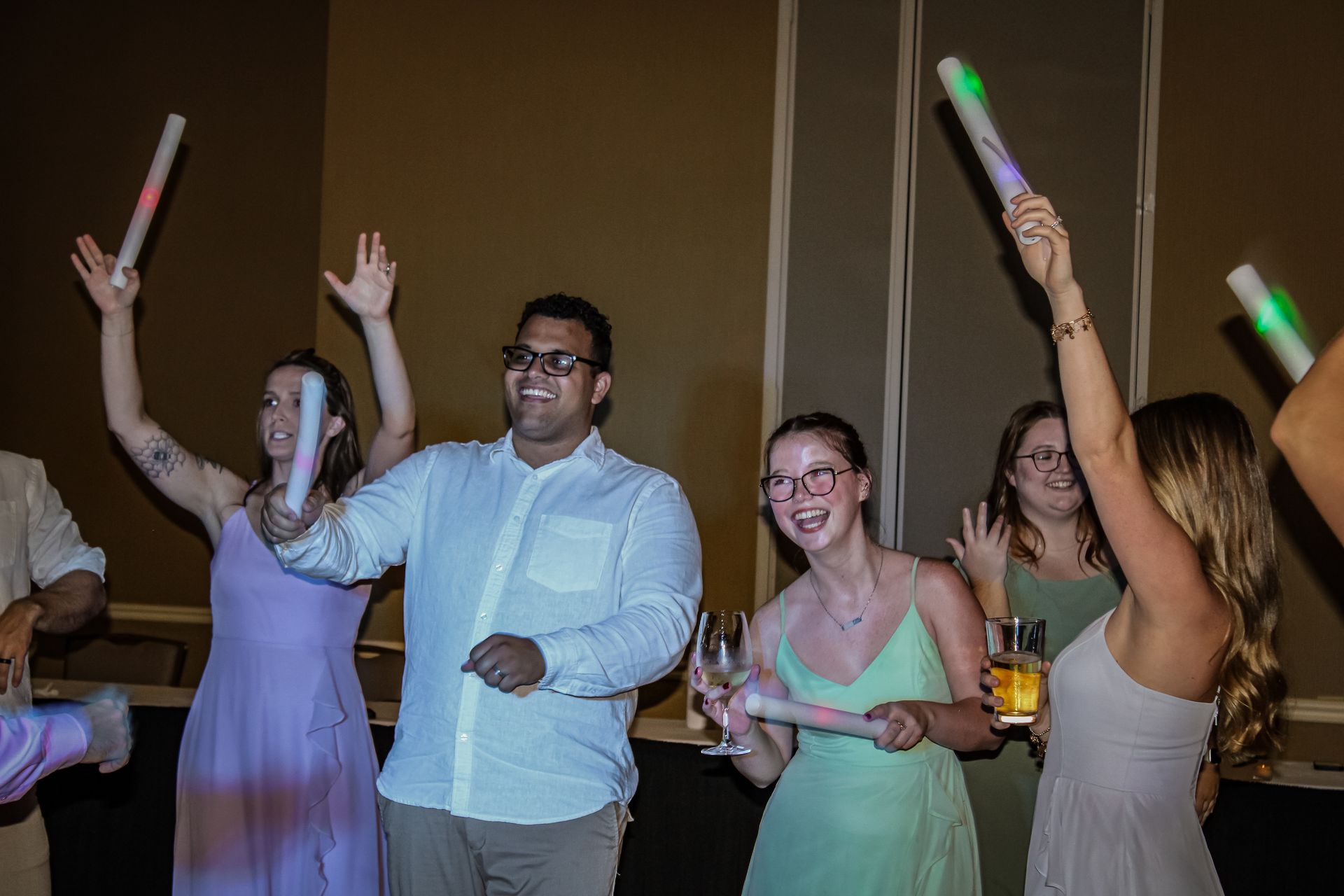 People dancing with glow sticks, arms raised. Party setting, wearing light-colored dresses and shirts.