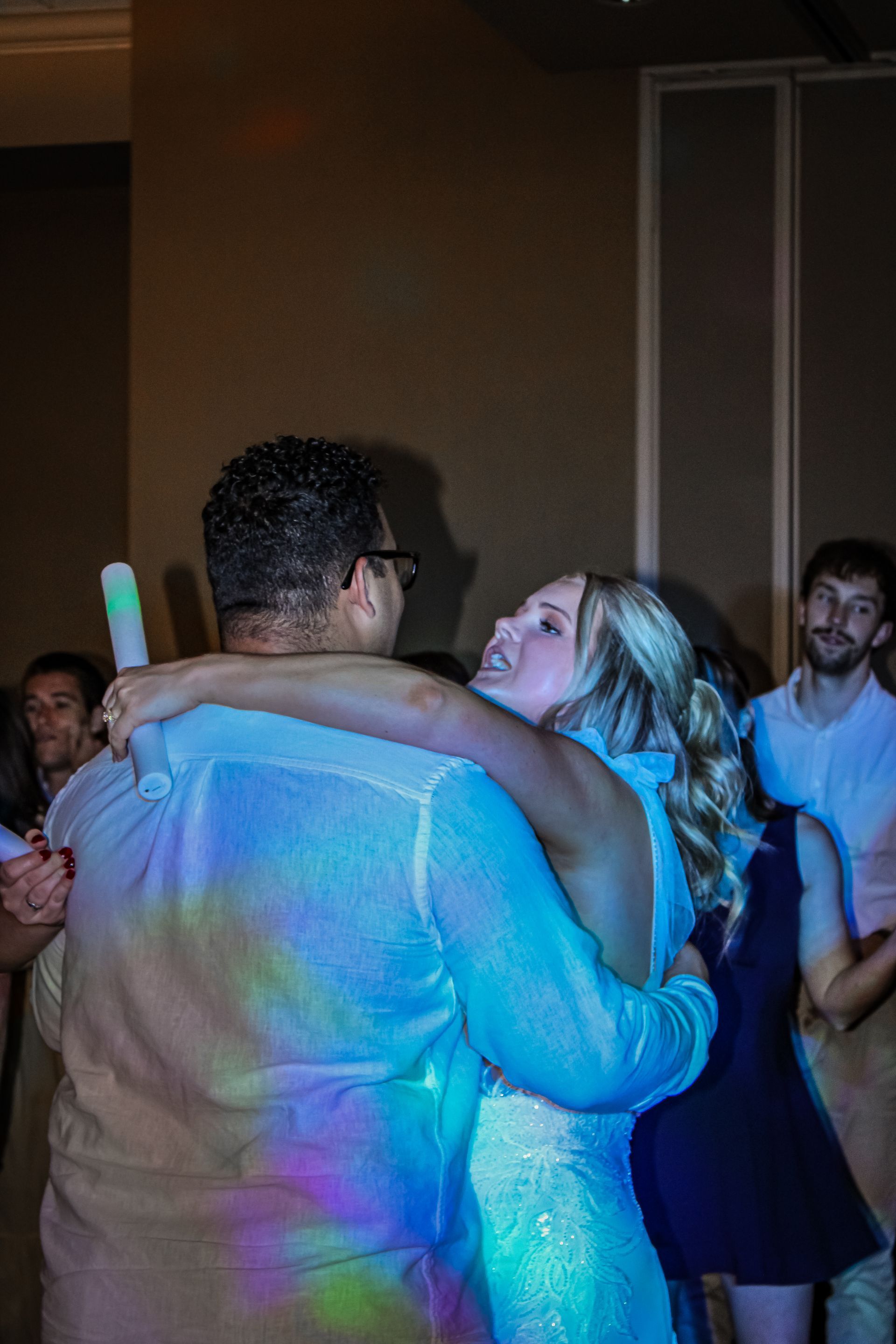 Couple dancing, embraced; colorful light illuminates them at a party, other guests nearby.