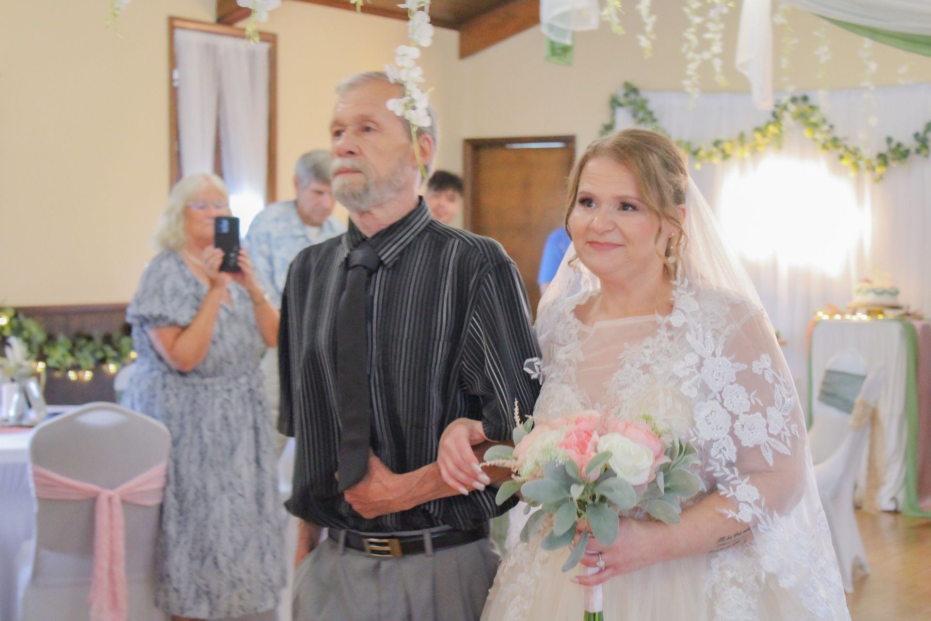 Bride walks down the aisle with her father, holding a bouquet. Reception hall with decorations.