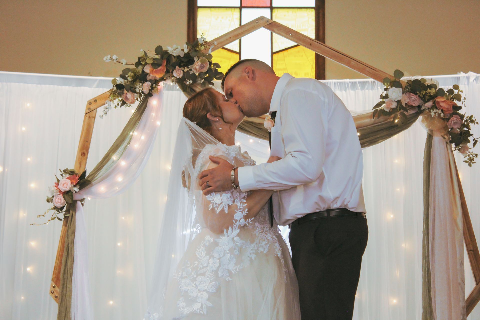 Couple kissing under a wooden arch decorated with flowers and fabric during a wedding ceremony.