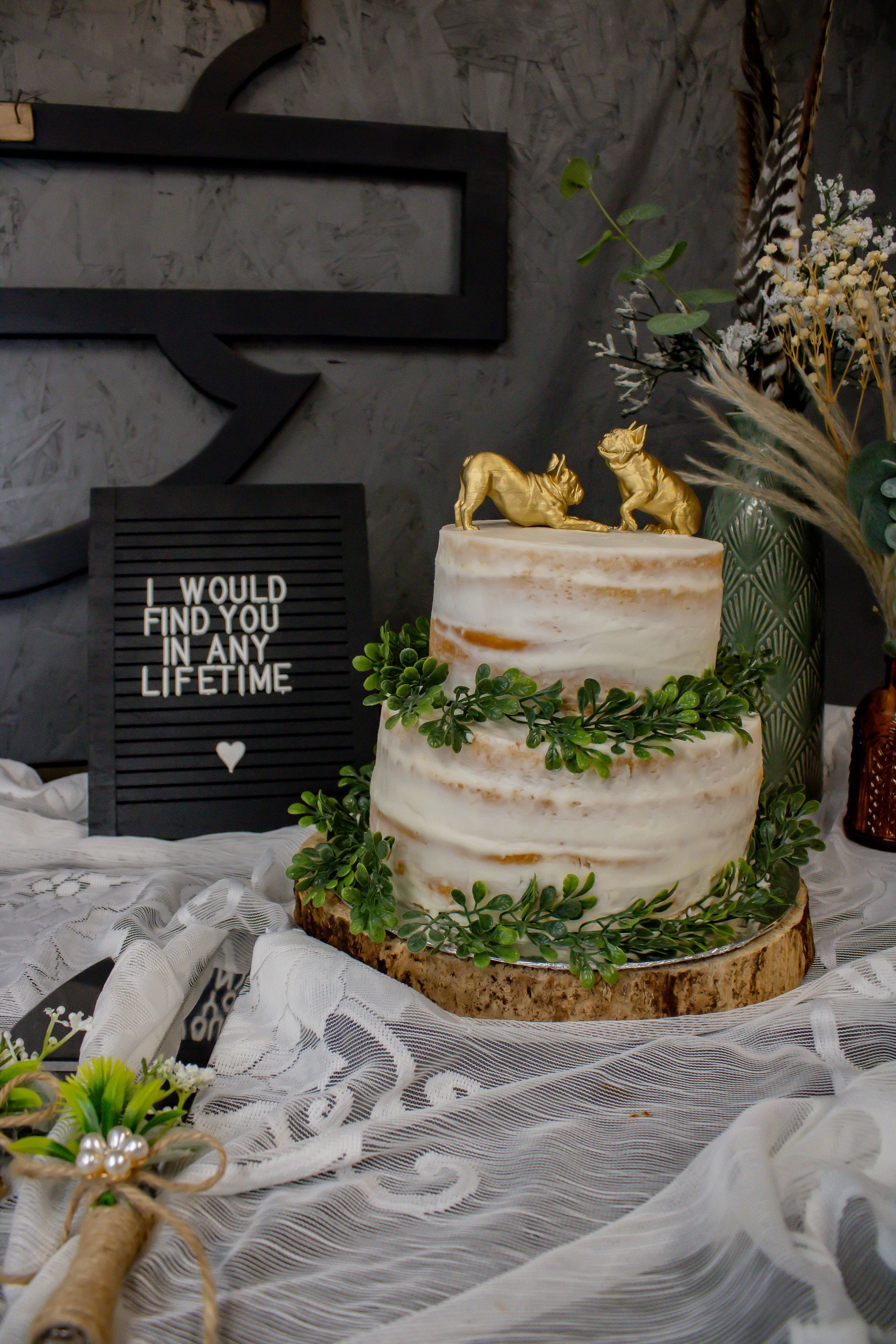 Two-tiered wedding cake with gold animal toppers and greenery, with a sign: