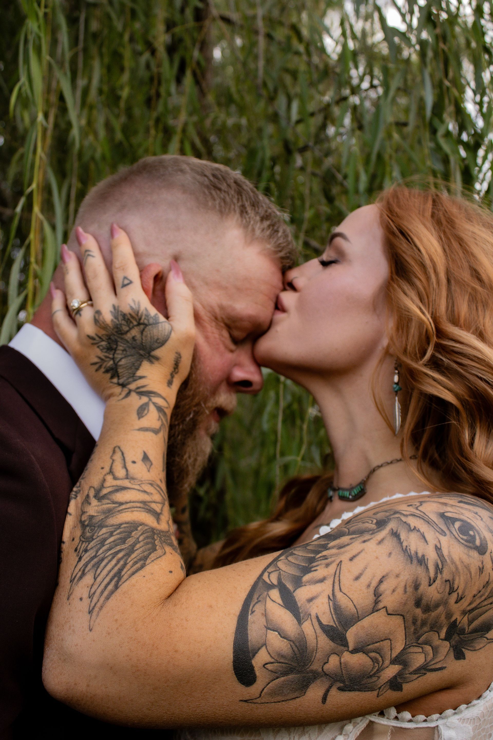 Bride kisses groom's forehead. Both have tattoos. Outdoors, against blurred foliage.