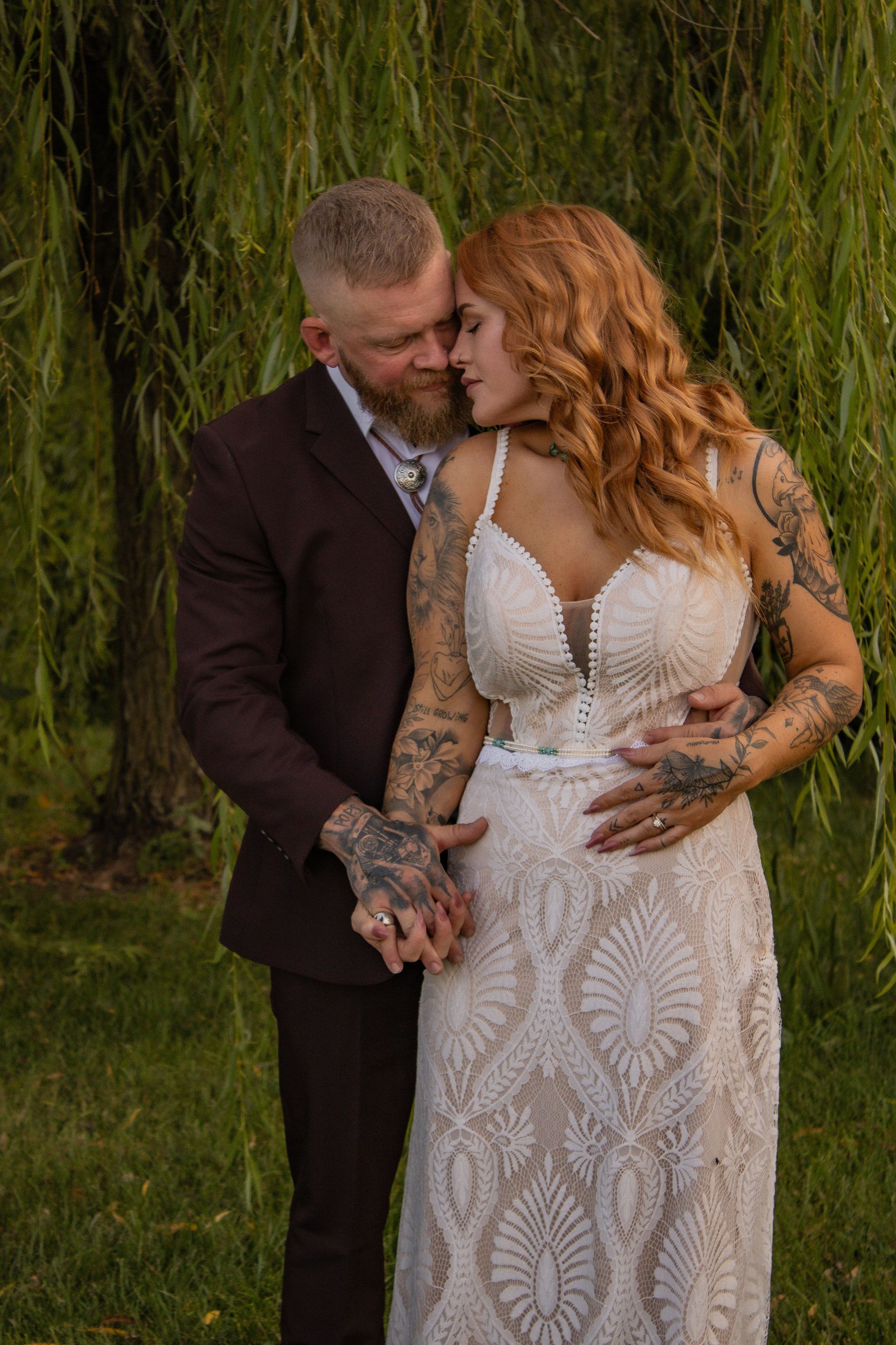 Bride and groom embrace outdoors, under weeping willow. Bride in white lace dress, groom in maroon suit.