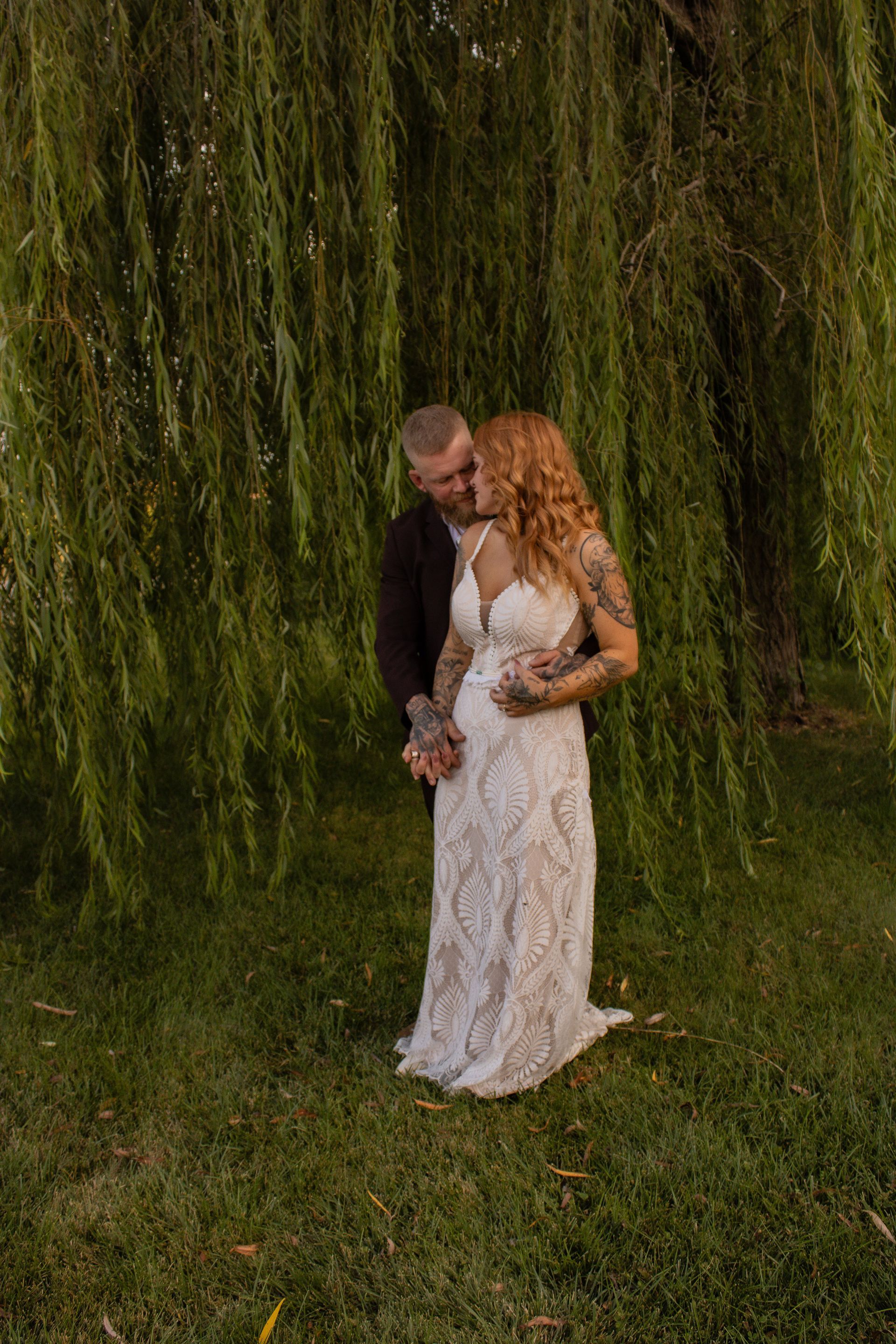 Couple embracing under a weeping willow tree; woman in a white lace dress and man in a black jacket, kissing.