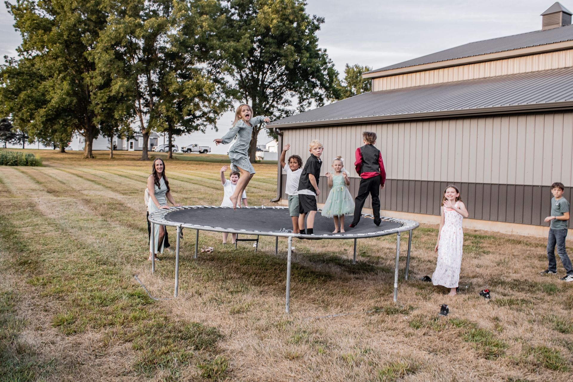 Children jumping on a trampoline in a field, a barn in the background.