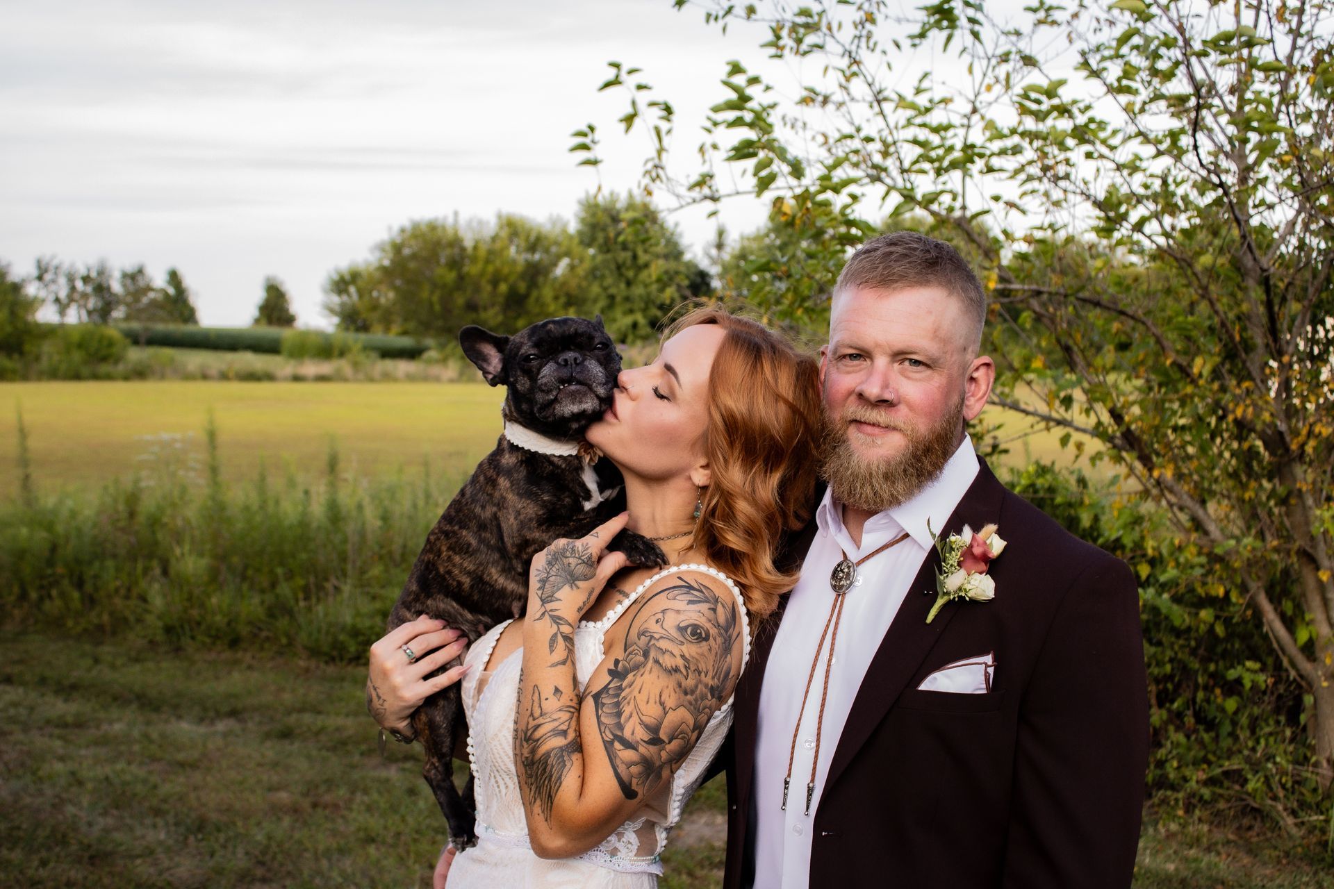 Couple in wedding attire with a dog; the woman kisses the dog while the man looks at the camera in a field.