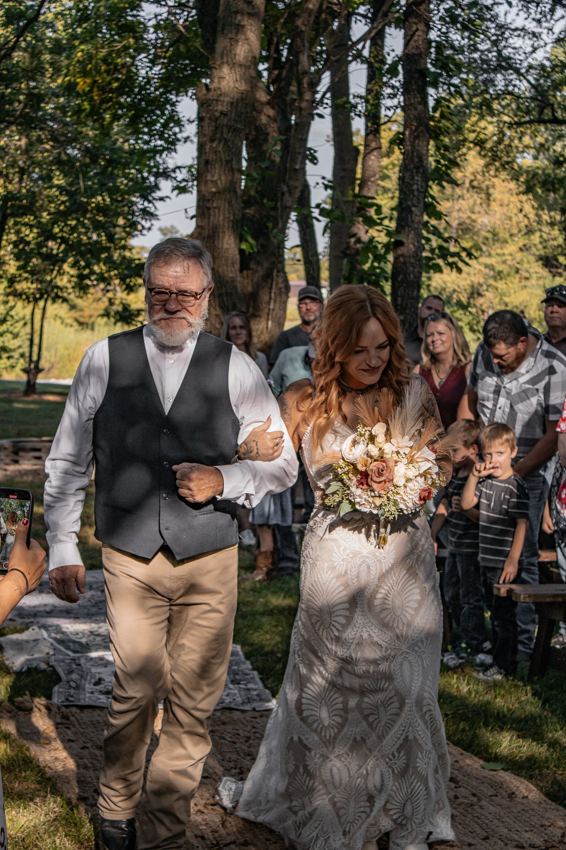 Bride and her father walking down an aisle, surrounded by guests, outdoor setting.