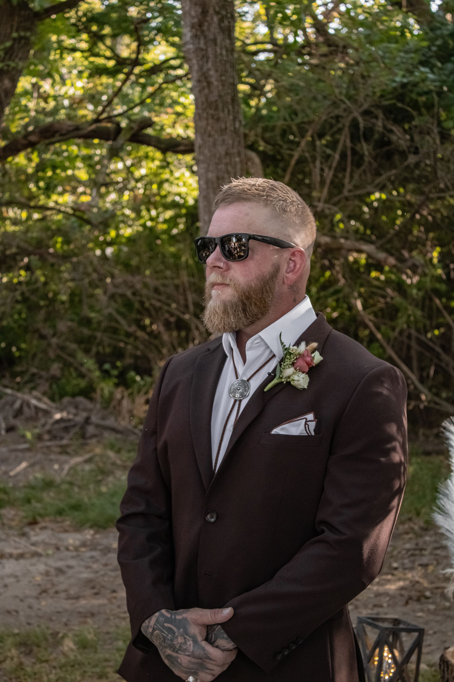 Man in brown suit and sunglasses, looking to the side, wearing a bolo tie and holding hands.