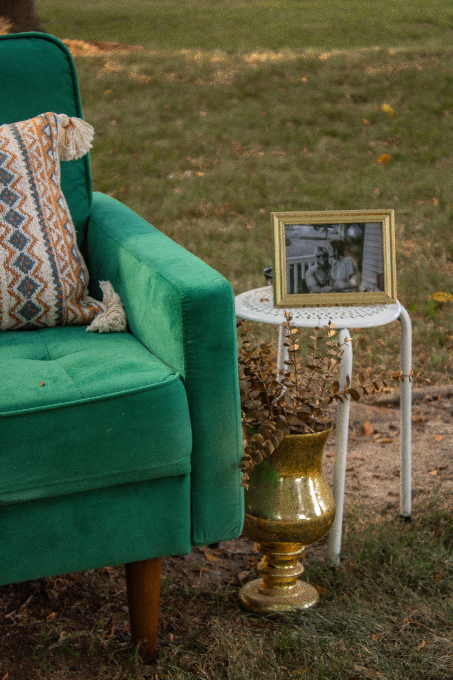 Emerald green armchair, pillow, small table with framed photo, gold vase, outdoors on grass.