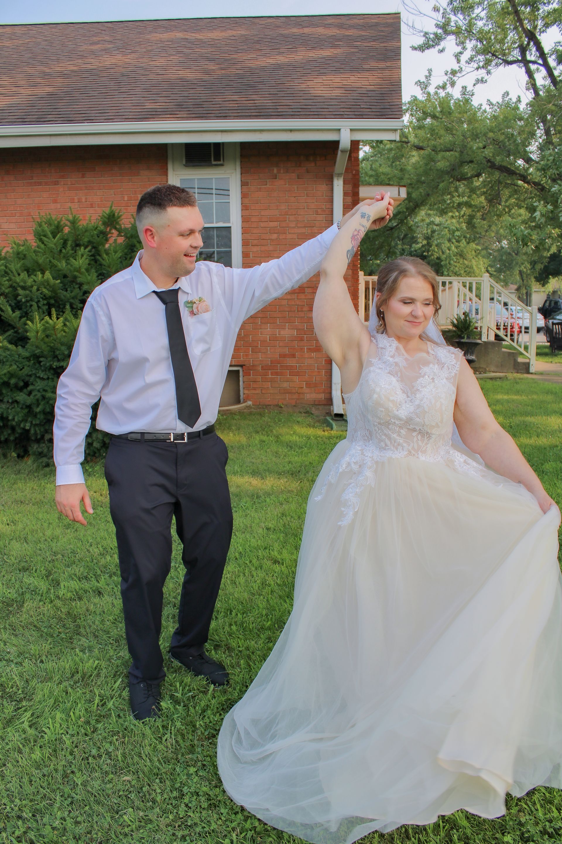 Wedding couple dancing in a yard. Bride in white gown, groom in black pants and tie, smiling. Green grass, brick building.