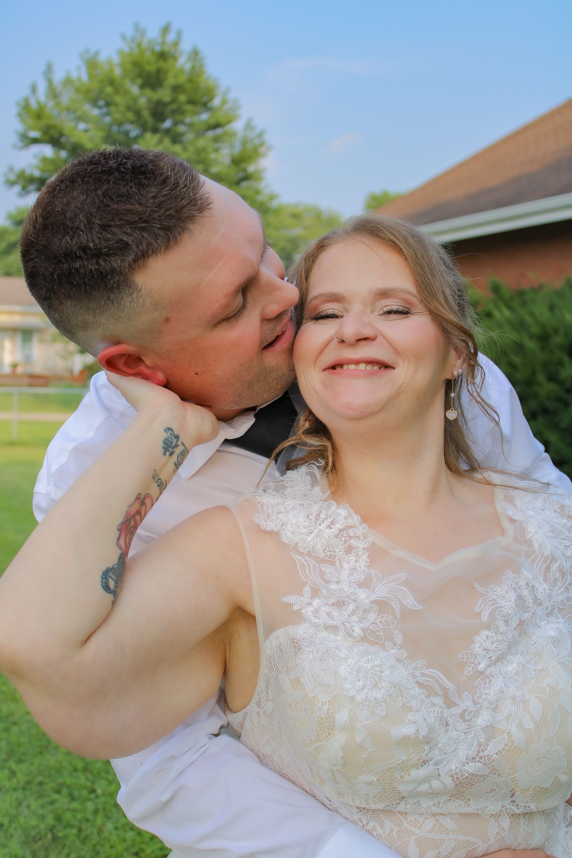 Couple embraces, woman in wedding dress smiles, man kisses her cheek, outdoors.