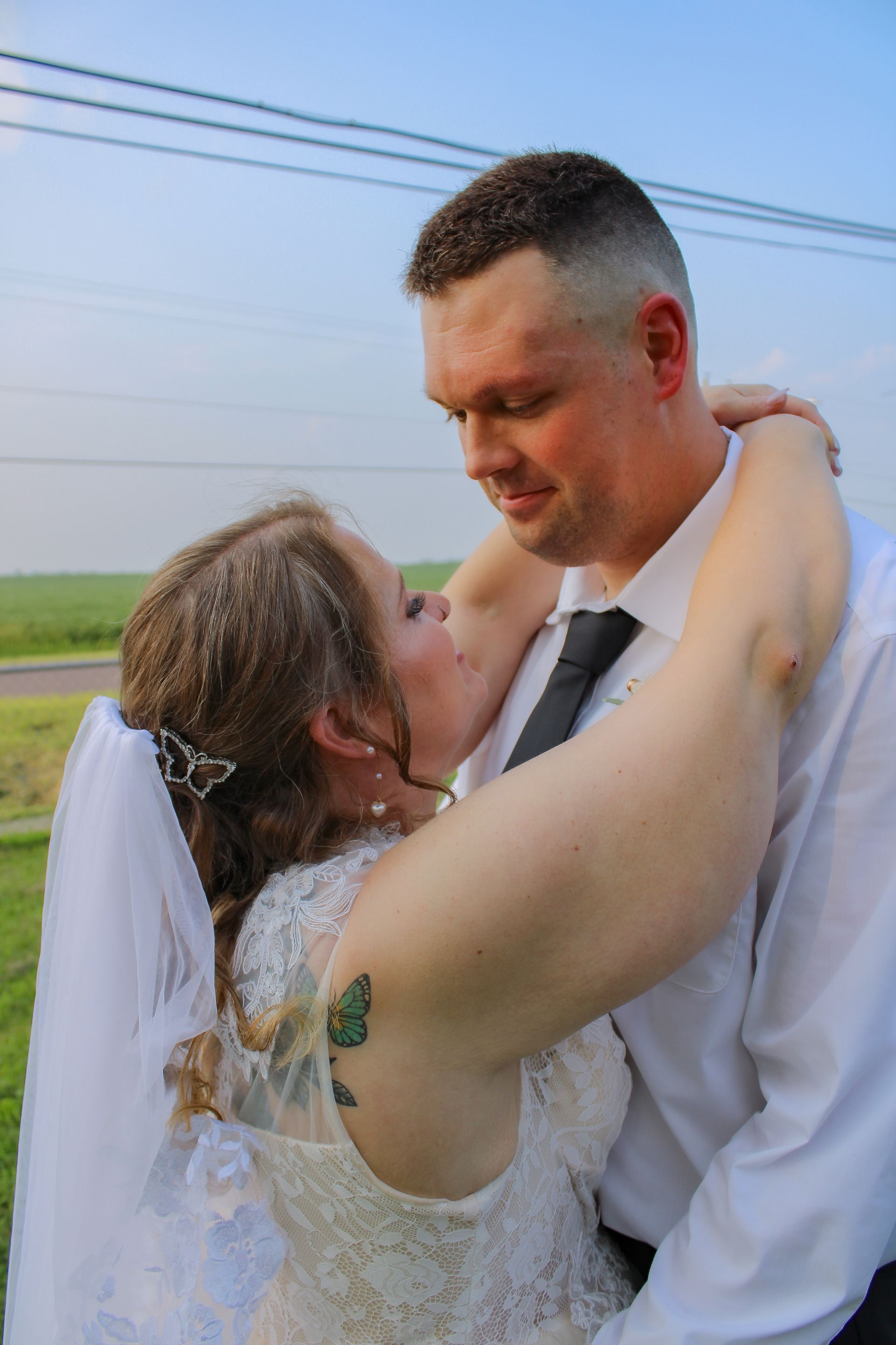 Wedding couple embracing outdoors; woman in white dress, veil, butterfly tattoo; man in suit.