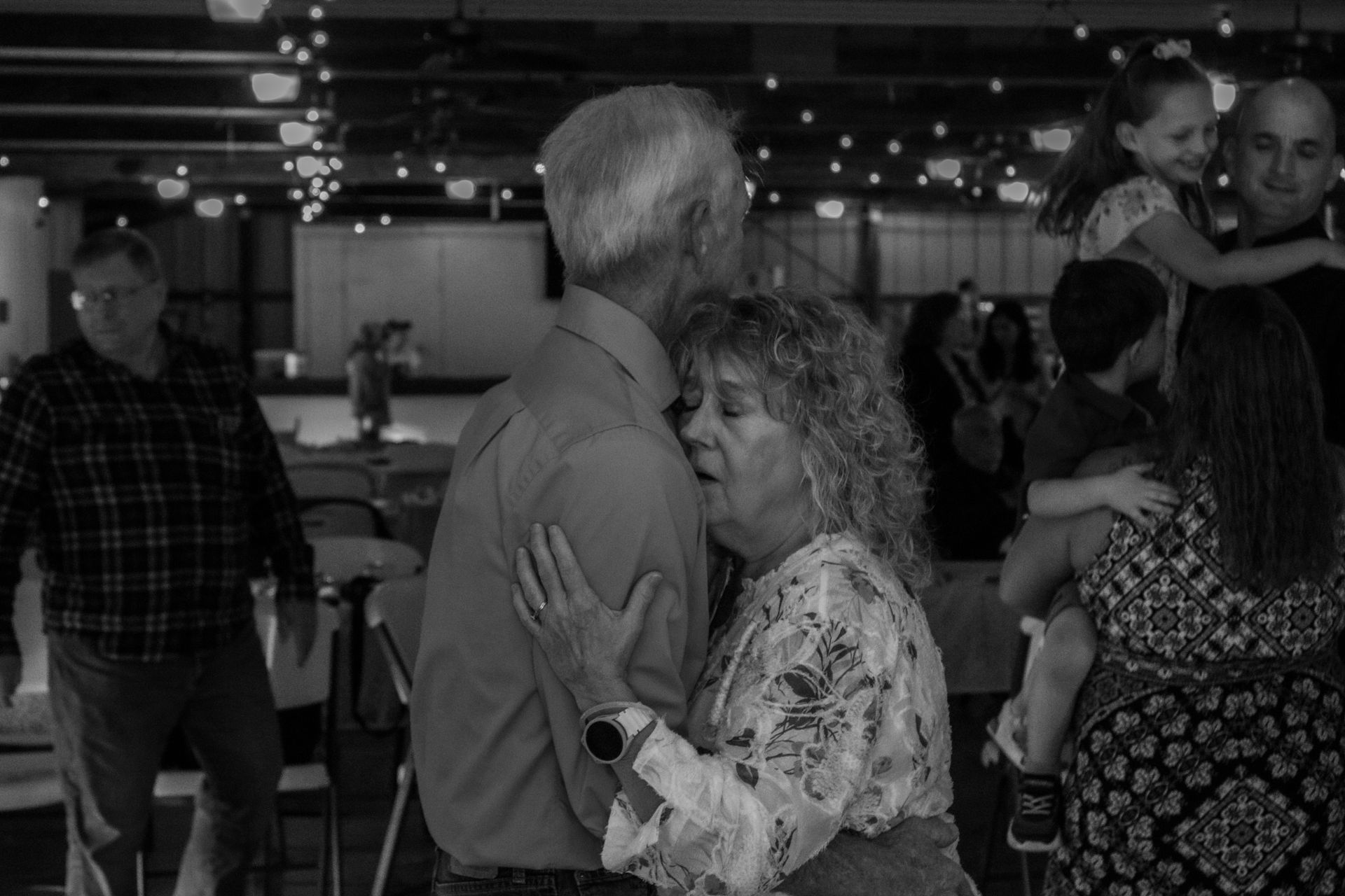 Couple slow dancing; others in the background. Woman’s head on man’s shoulder. String lights and general celebration setting.