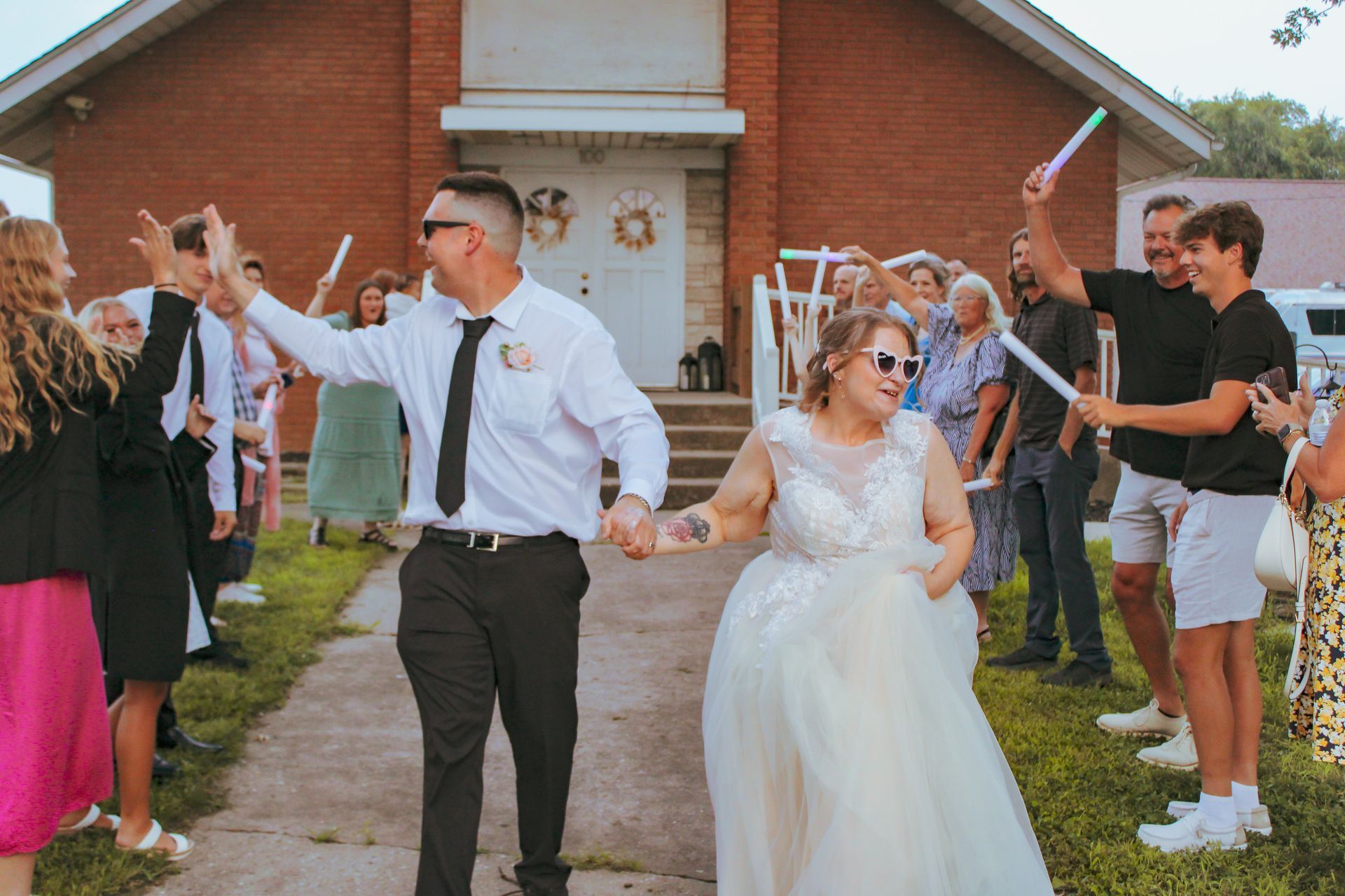 Newlyweds exit a church, hand in hand, cheered by guests waving glow sticks.