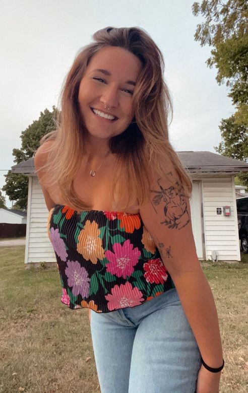 A woman wearing a floral strapless top and jeans is standing in front of a house.