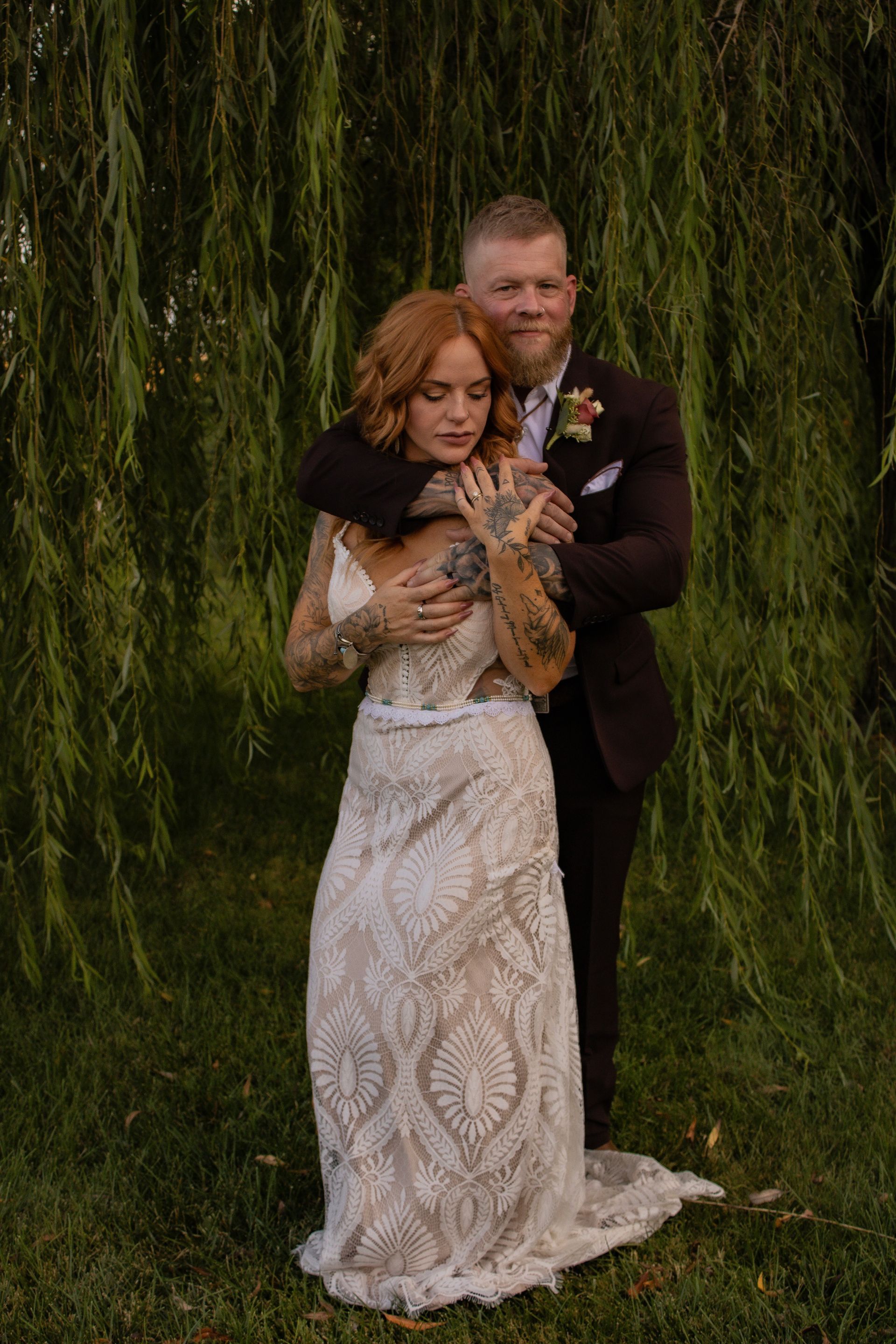 Bride in lace dress embraced by groom in brown suit, under weeping willow.