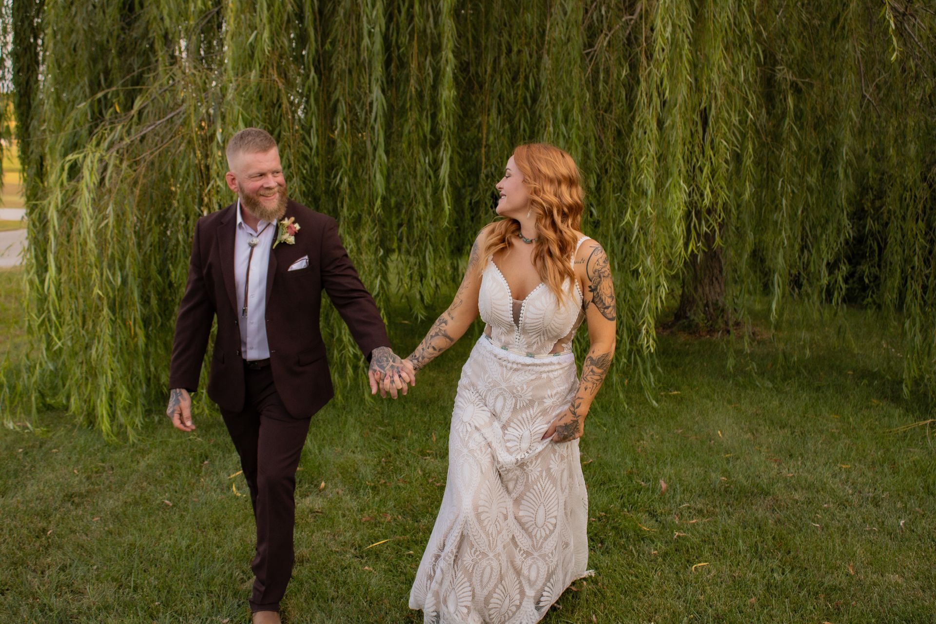 Couple holding hands, smiling, walking in front of weeping willow tree; It's their wedding day!