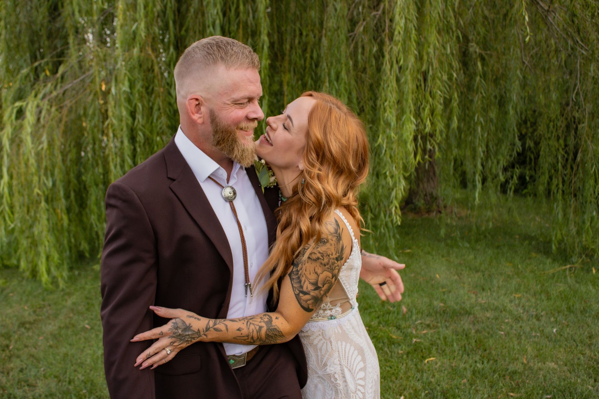 Wedding couple embracing outdoors; woman kisses man's beard, both smiling. Green trees in the background.