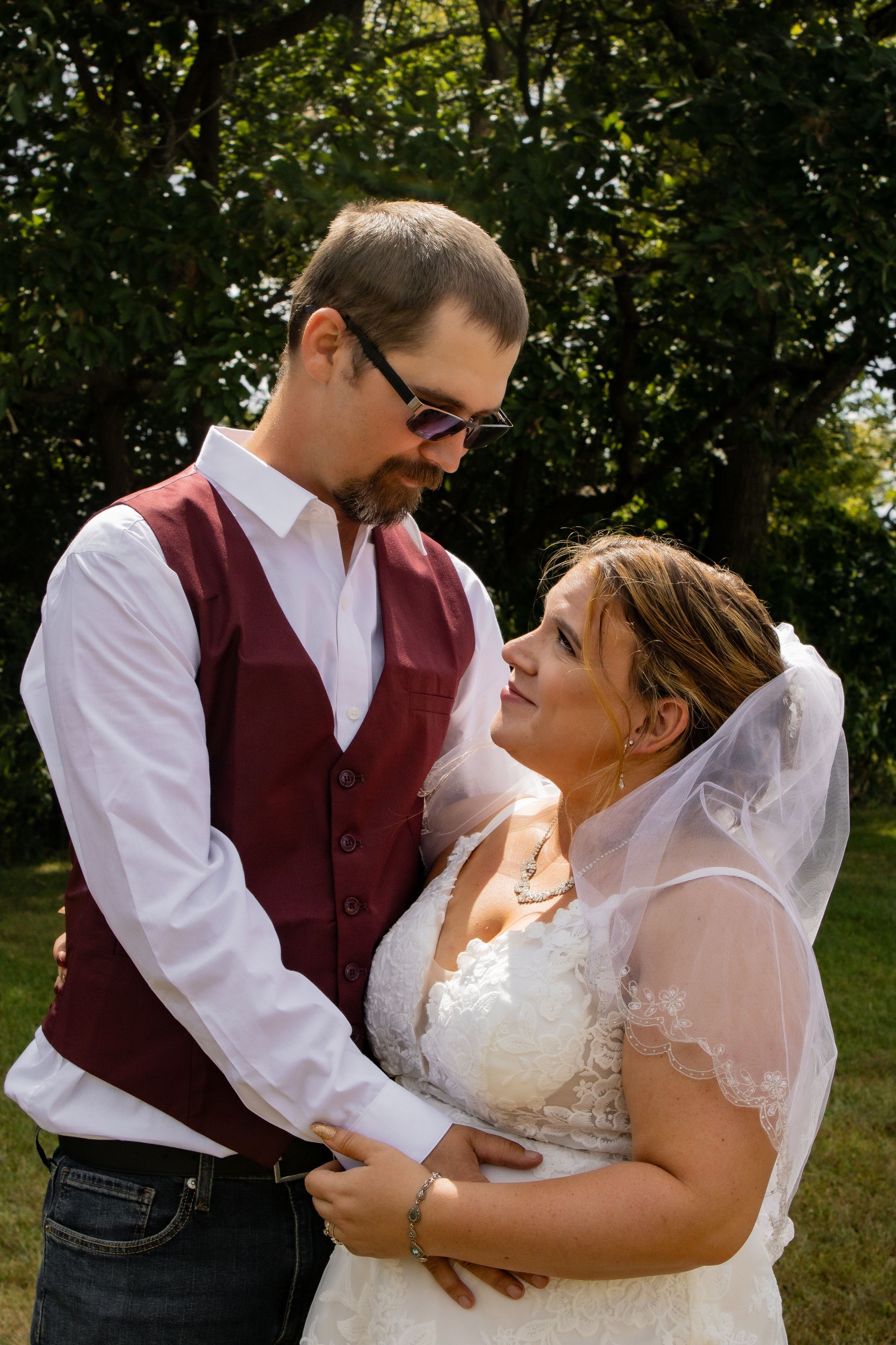 Couple gazing at each other, bride in white dress and veil, groom in vest and sunglasses, outdoors.
