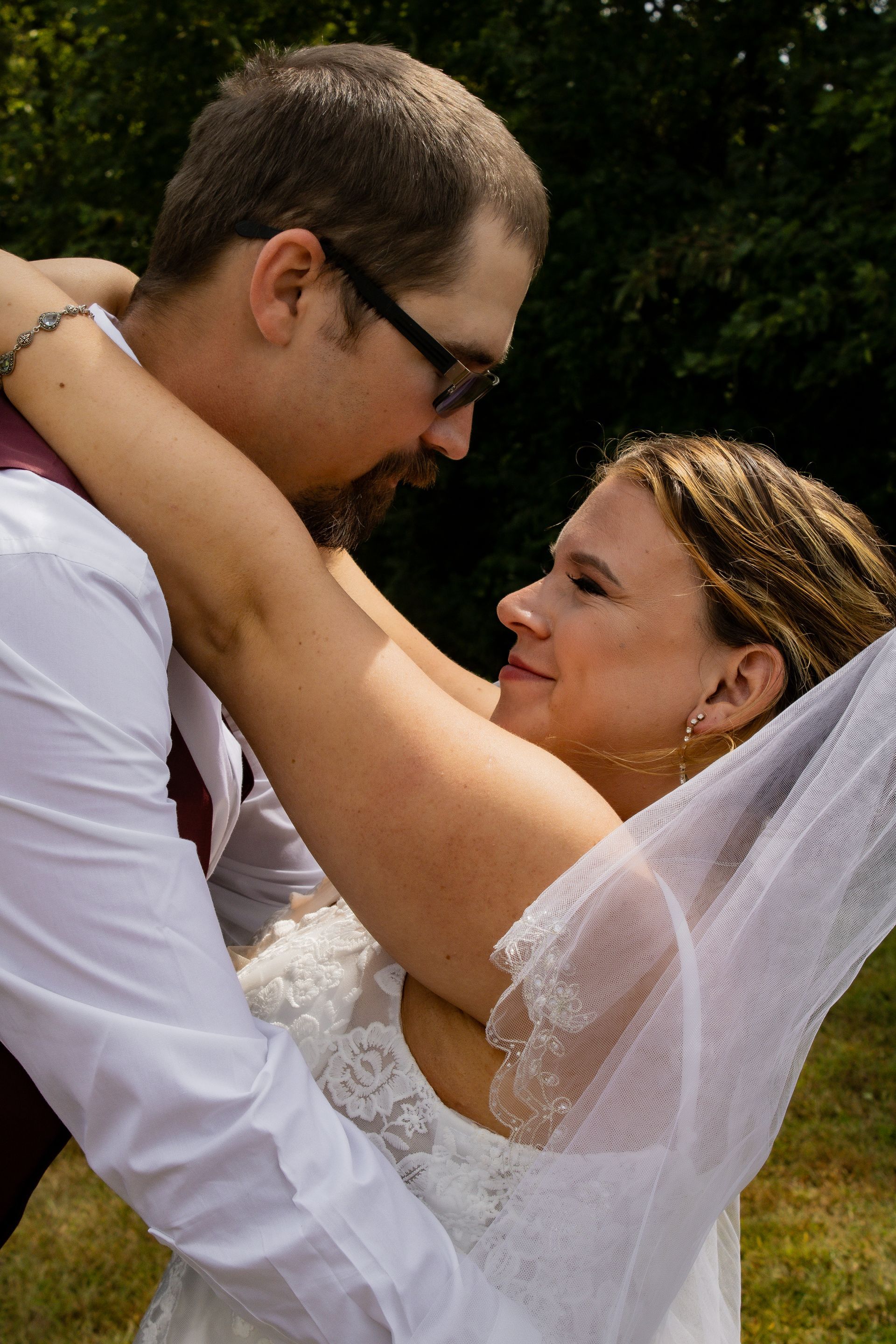 Couple embraces, bride in white dress, groom in vest, looking lovingly at each other, outdoors.