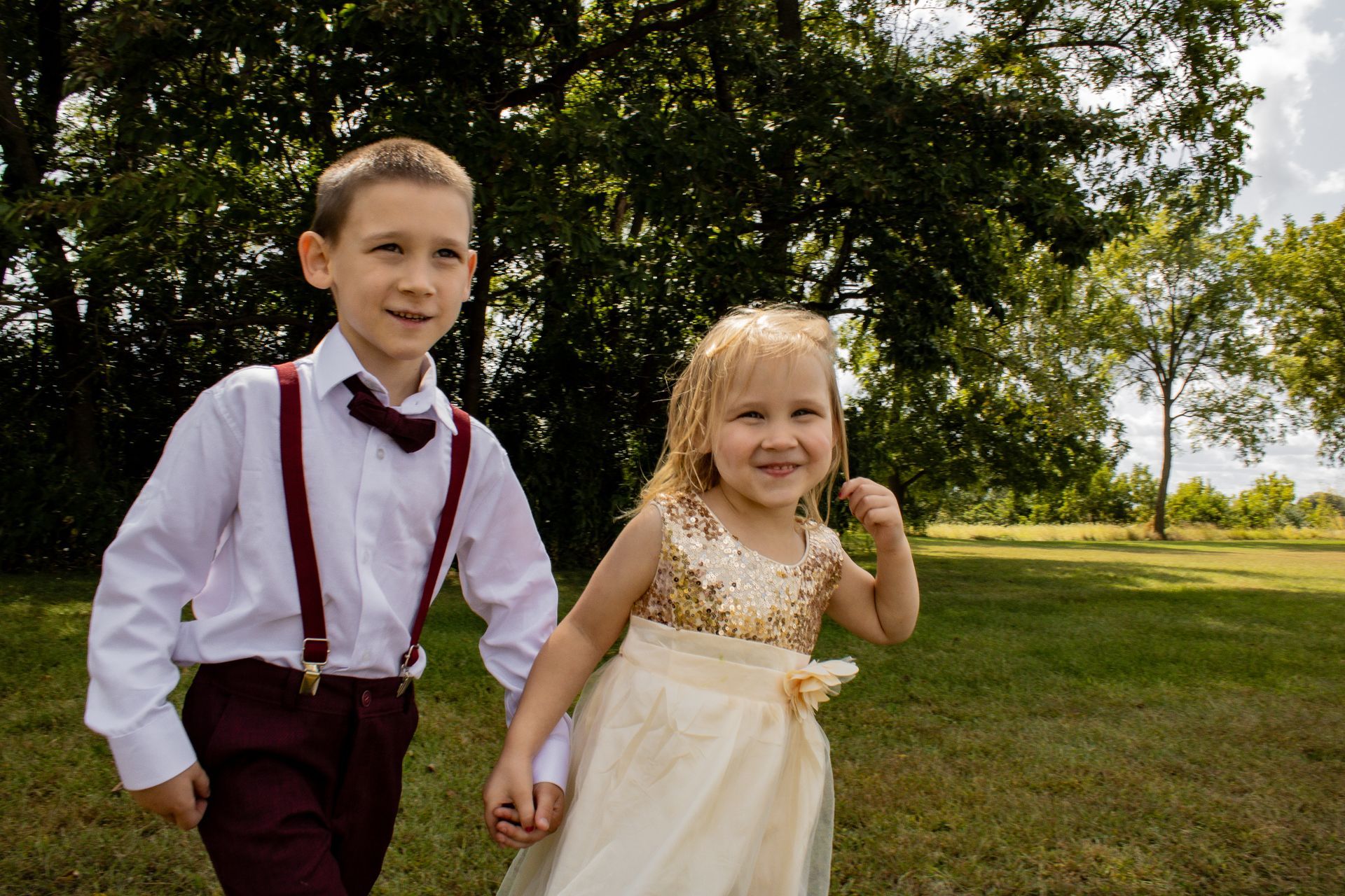 Boy and girl in formal attire holding hands, smiling while walking through a grassy area near trees.