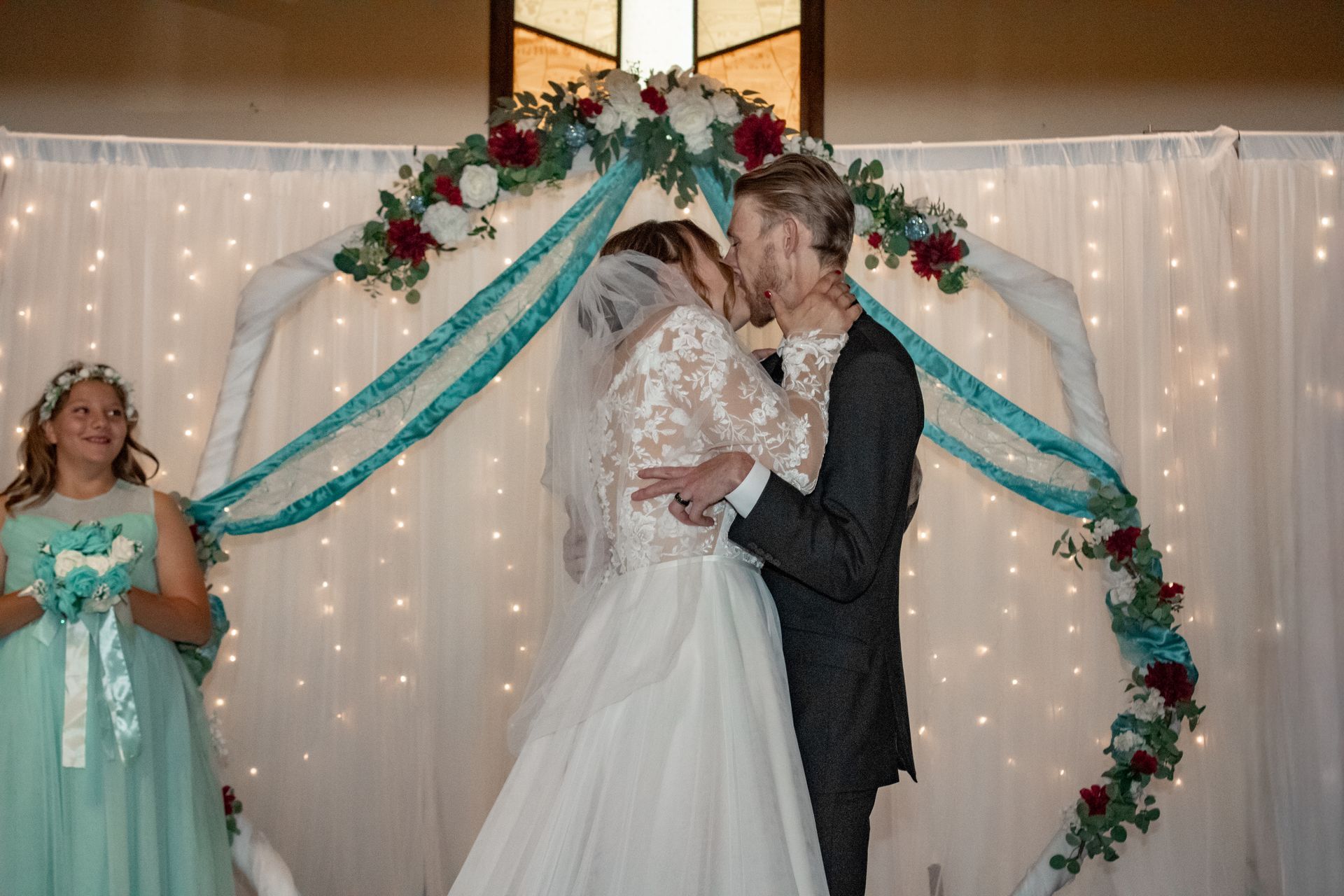 Couple kissing under decorated arch during wedding ceremony, bridesmaid watches.