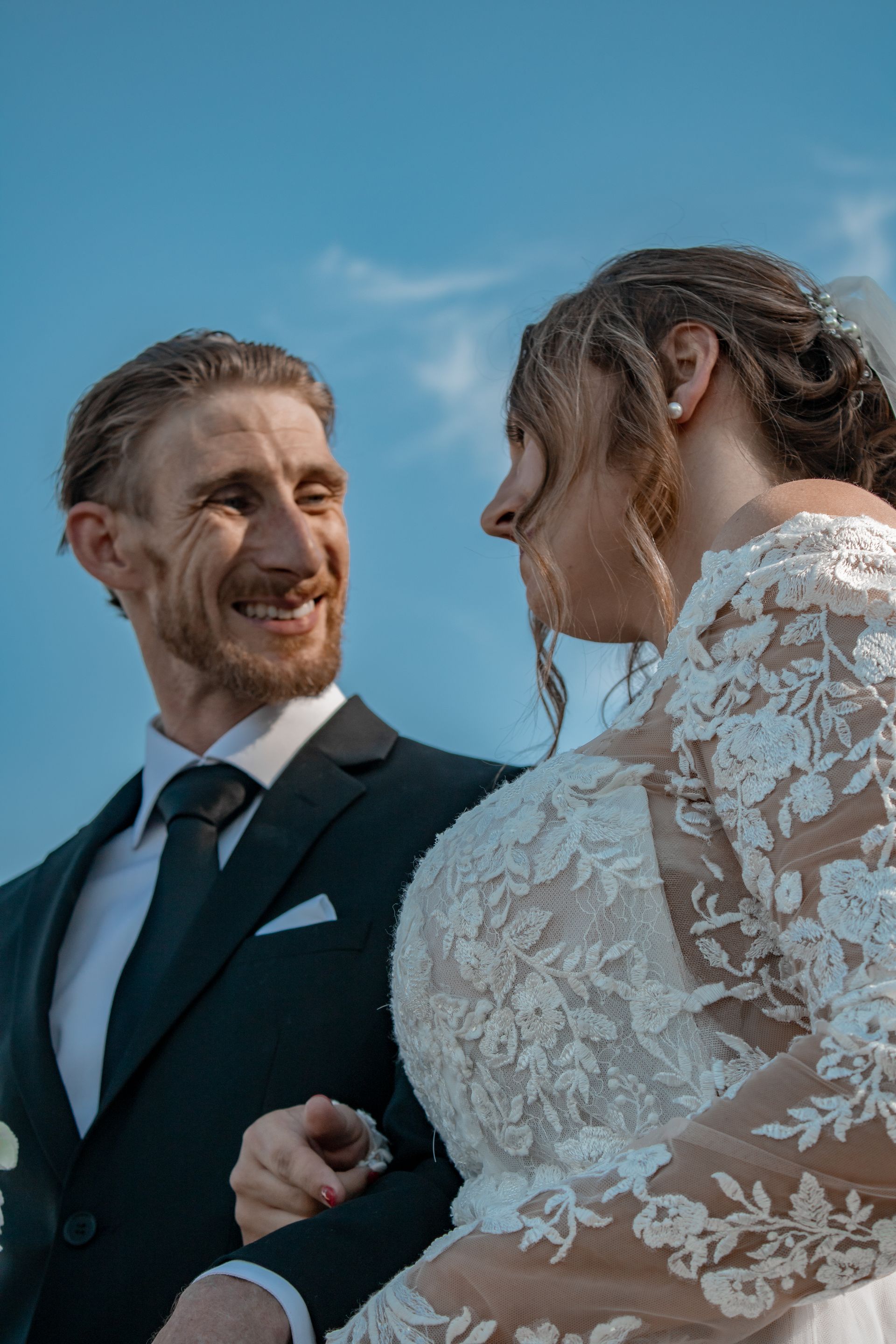 Groom in suit smiles at bride in white lace dress under blue sky, wedding day.