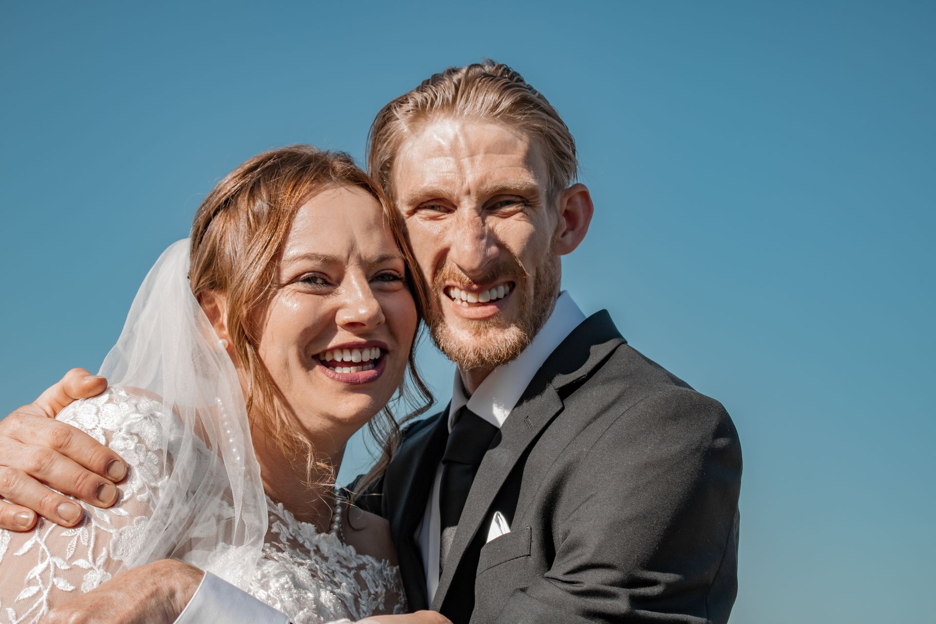 Bride and groom smiling, embracing outdoors. Bride wears a white dress and veil, groom in a dark suit. Blue sky.