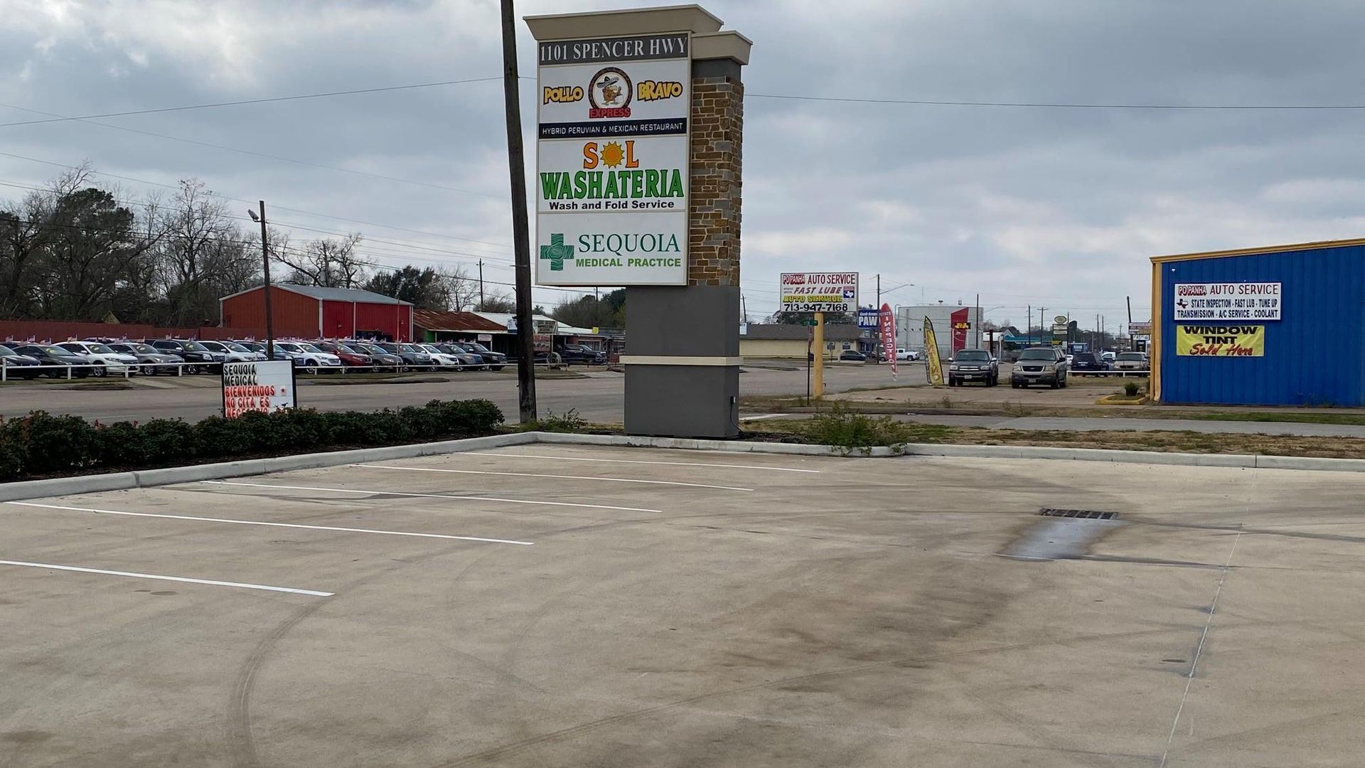 Shopping center with sign listing businesses, parking lot, overcast sky.