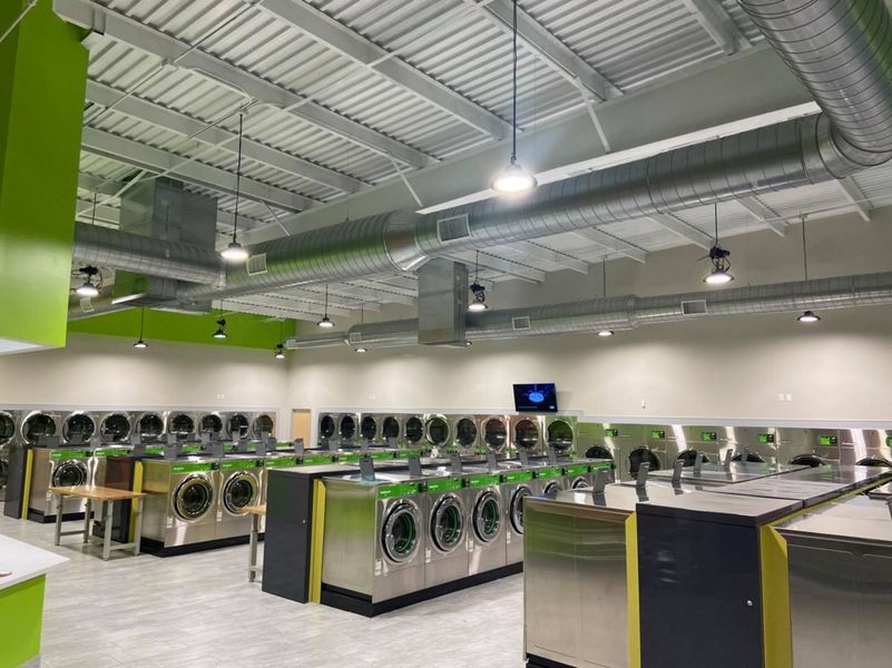 Interior of a brightly lit laundromat with rows of washers and dryers, green accents, and exposed ceiling ventilation.
