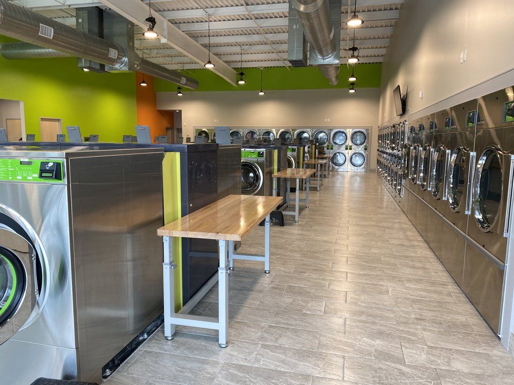 A well-lit laundromat with rows of stainless steel washing machines and dryers. Green and orange accent walls.