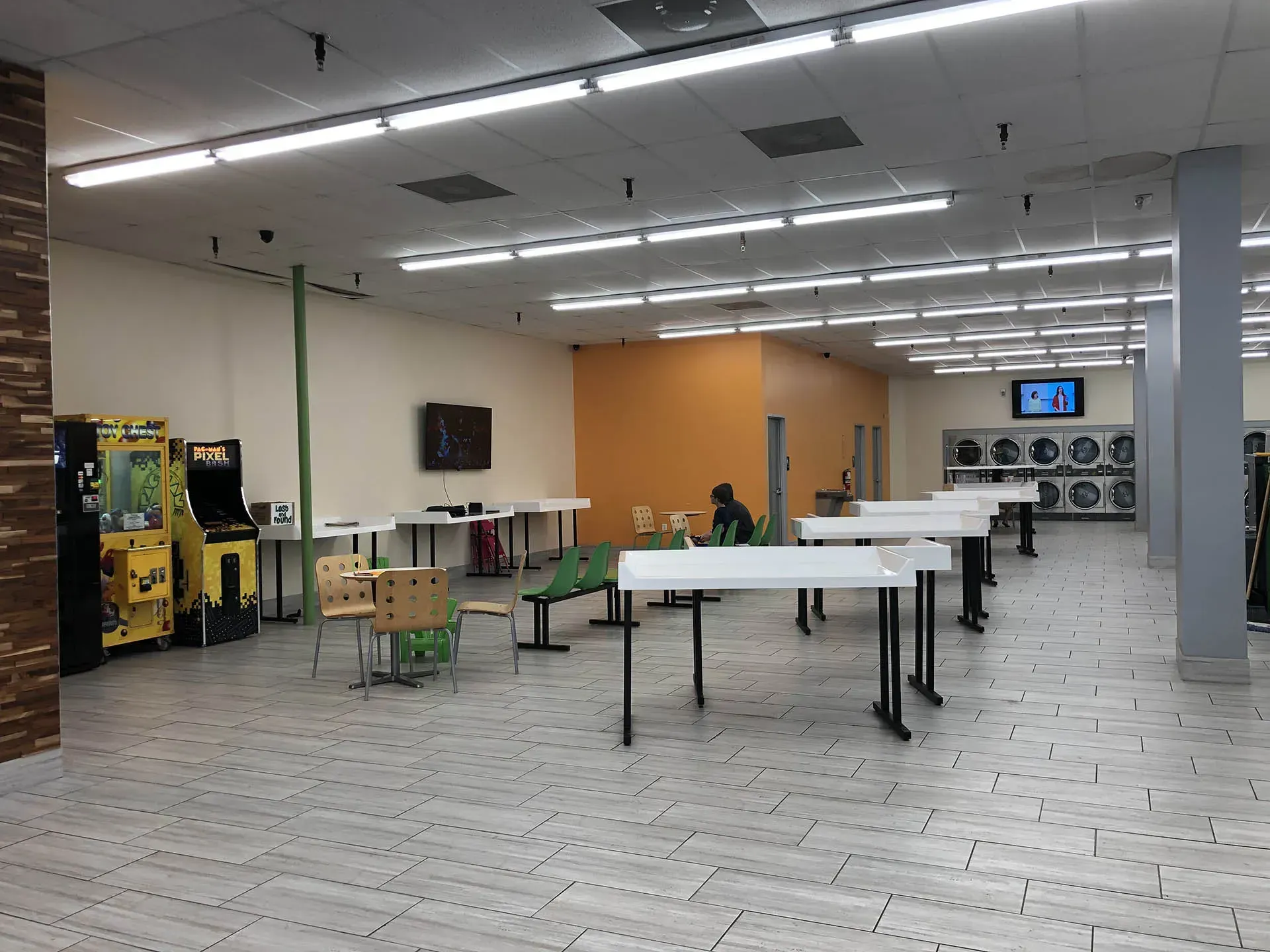Interior of a laundromat with tables, arcade games, and washing machines. Light-colored walls and flooring.