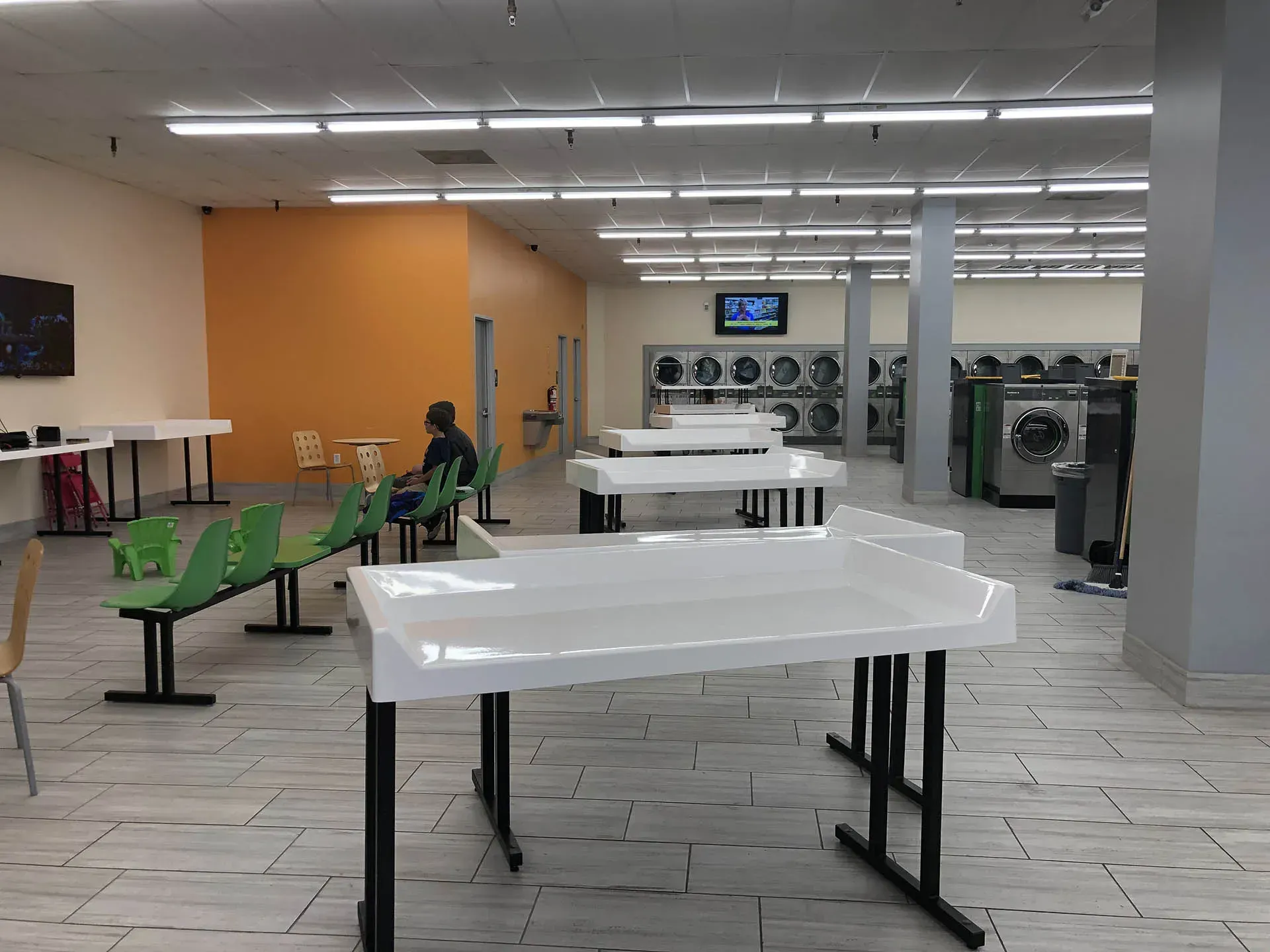 Laundry room with folding tables, seating, and washing machines. One person is sitting. Fluorescent lighting.