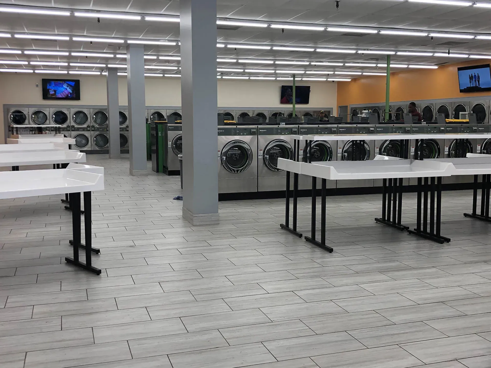 Interior of a laundromat with rows of washers and dryers, folding tables, and two TVs on the walls.