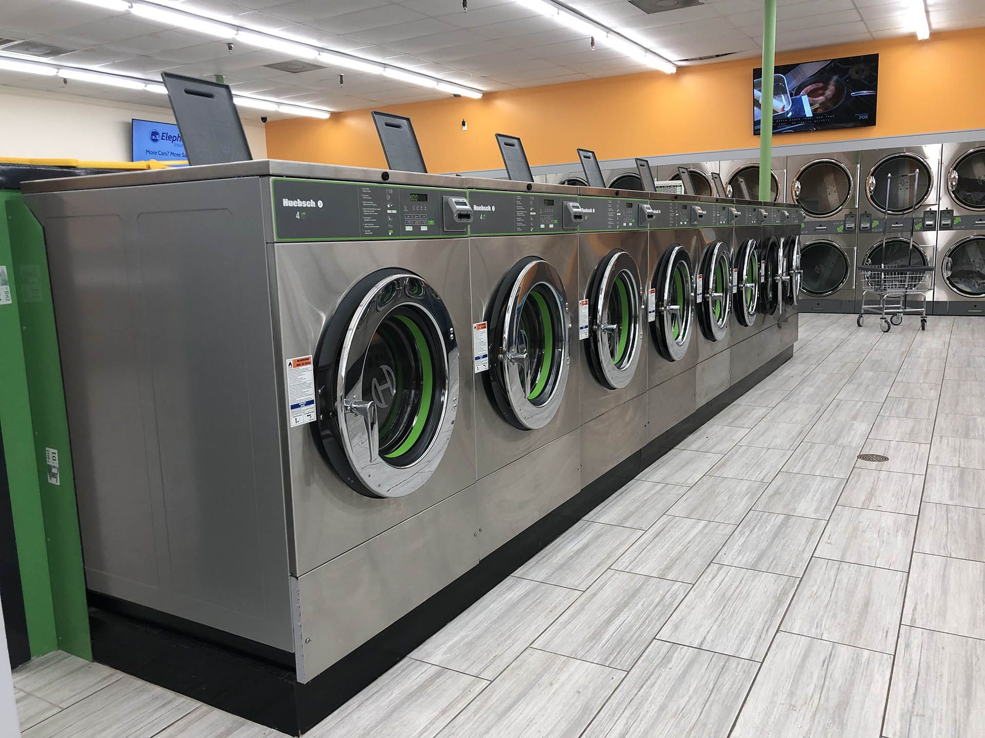 Row of washing machines in a laundromat, stainless steel with green accents. Light-colored tiled floor, orange and white walls.