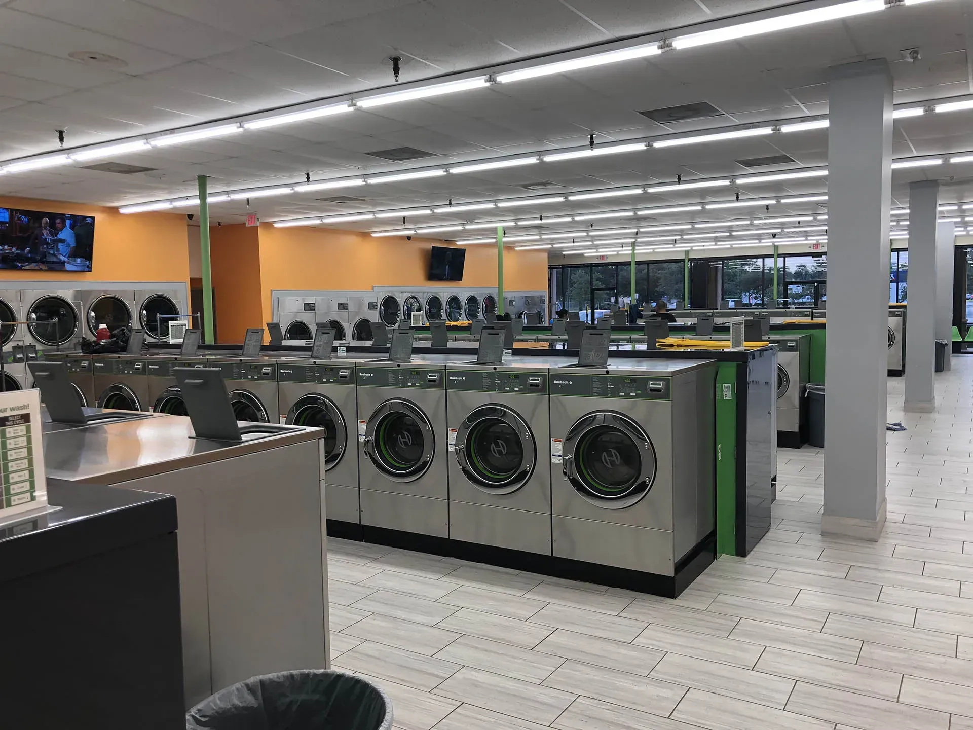 Inside a laundromat, washers and dryers in rows. Fluorescent lighting.