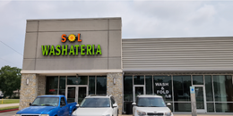 Exterior of Sol Washateria, a laundromat with a tan facade and large windows, with three vehicles parked outside.