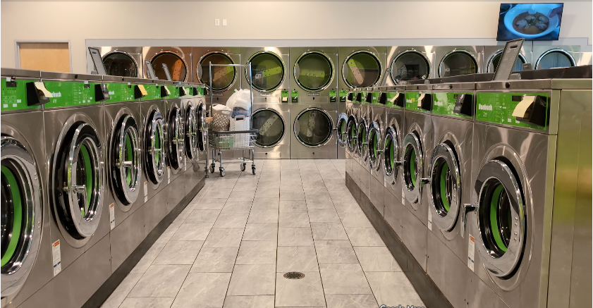 Rows of stainless steel washing machines in a laundromat, with green accents and large glass doors.