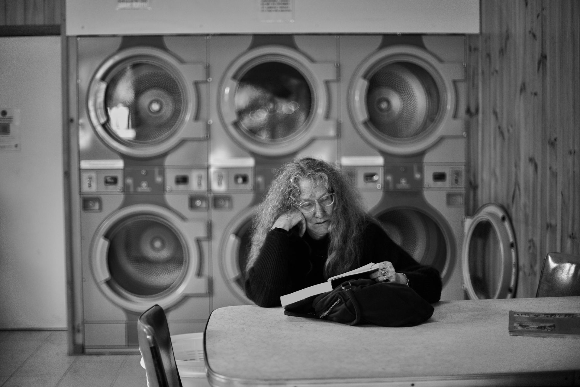Woman reads book in laundromat, leaning on folding table; washers in background. Black and white.