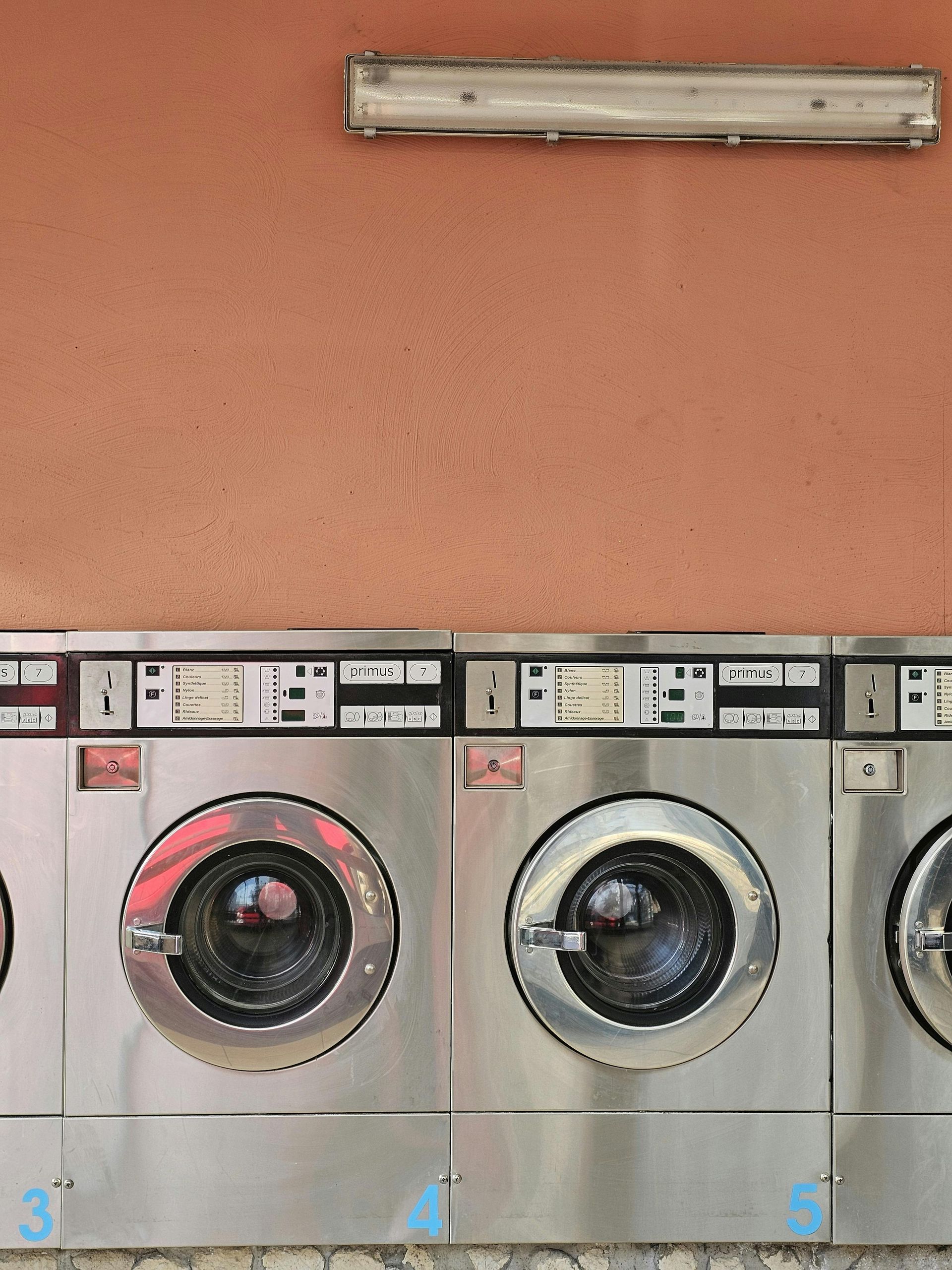 Row of silver washing machines against a peach-colored wall, with overhead fluorescent lighting.