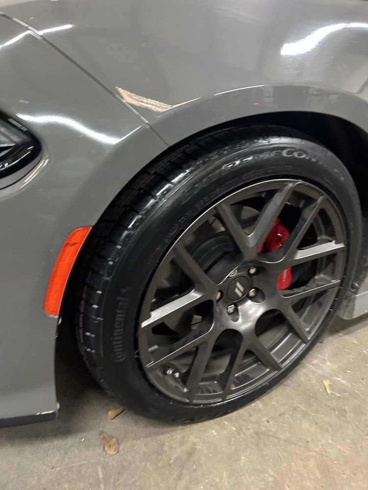 Gray car wheel with red brake caliper, set in front of a gray fender, on a garage floor.