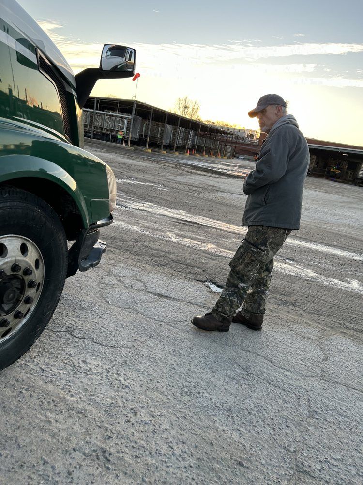 A trucker stands near his green semi-truck in a lot, leaning with arms crossed.