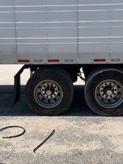 Semi-trailer's wheels and side. The silver trailer sits on a gravel surface. Two tire rims and tires are visible. 
