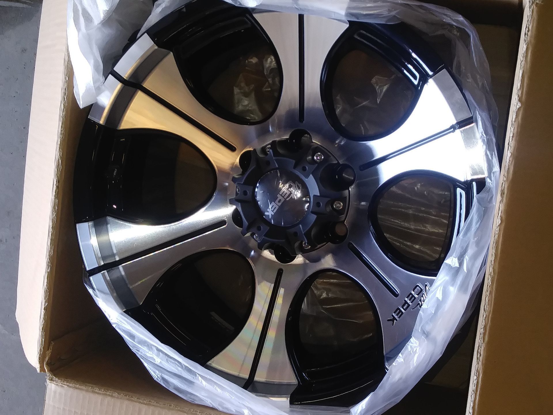 Black and silver car wheel in a cardboard box, wrapped in plastic.