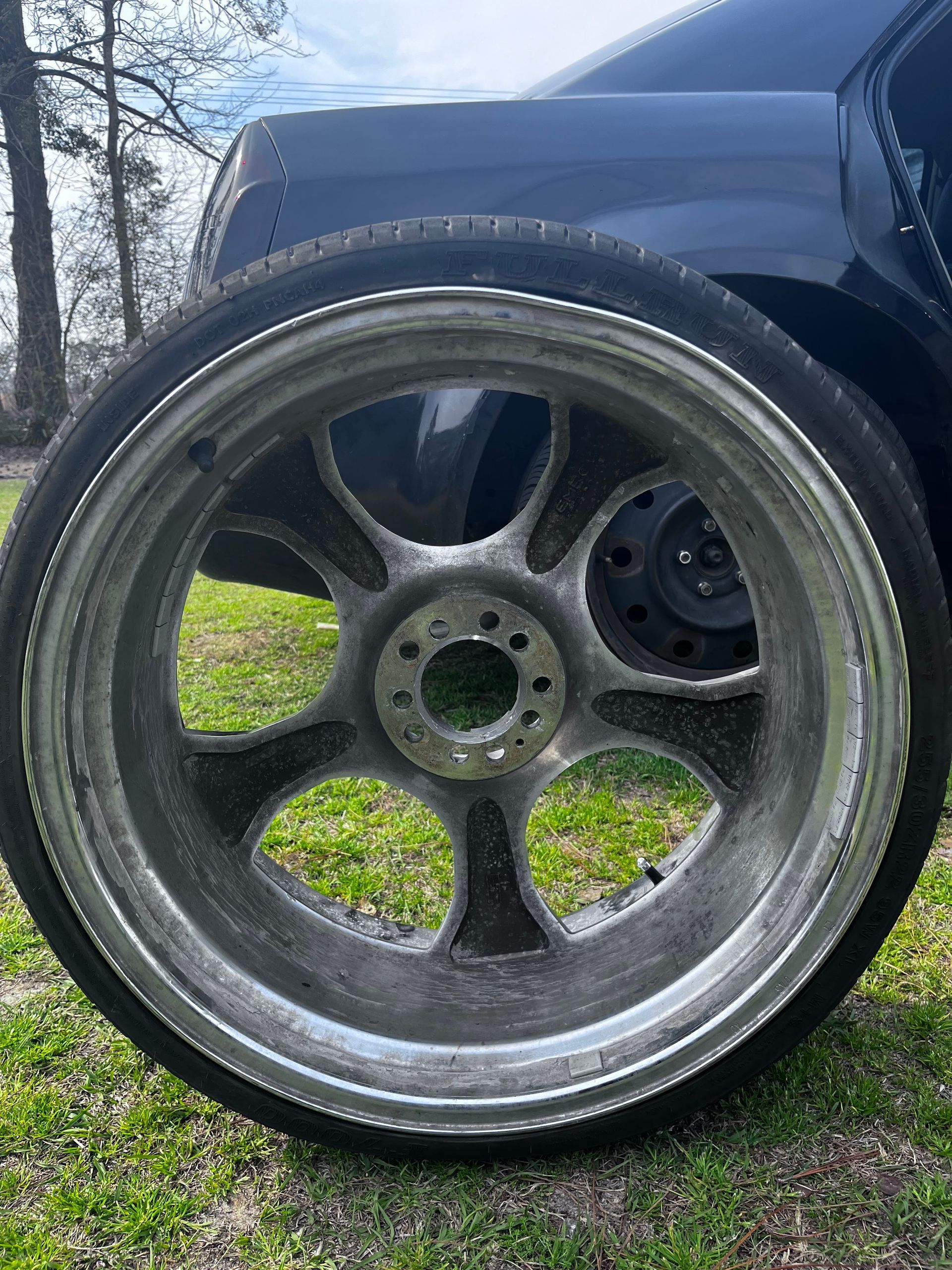 Silver alloy car wheel with a tire, sitting on green grass near a black car.