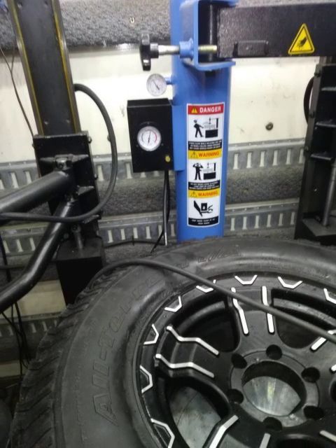 A tire on a black and silver rim being inflated by a machine, near blue equipment with danger warnings.