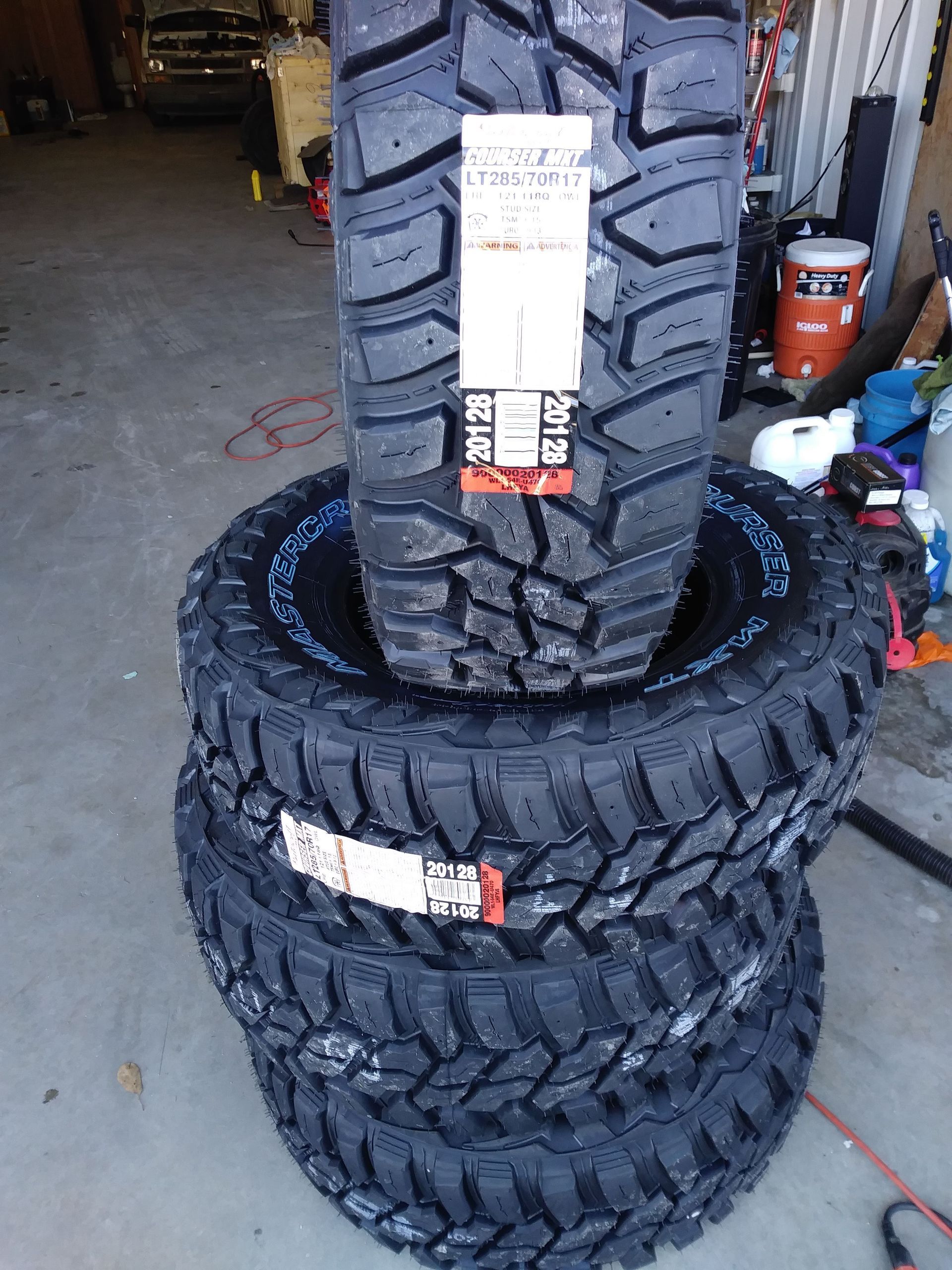 Four black off-road tires stacked on a concrete floor, with a tire label on each.